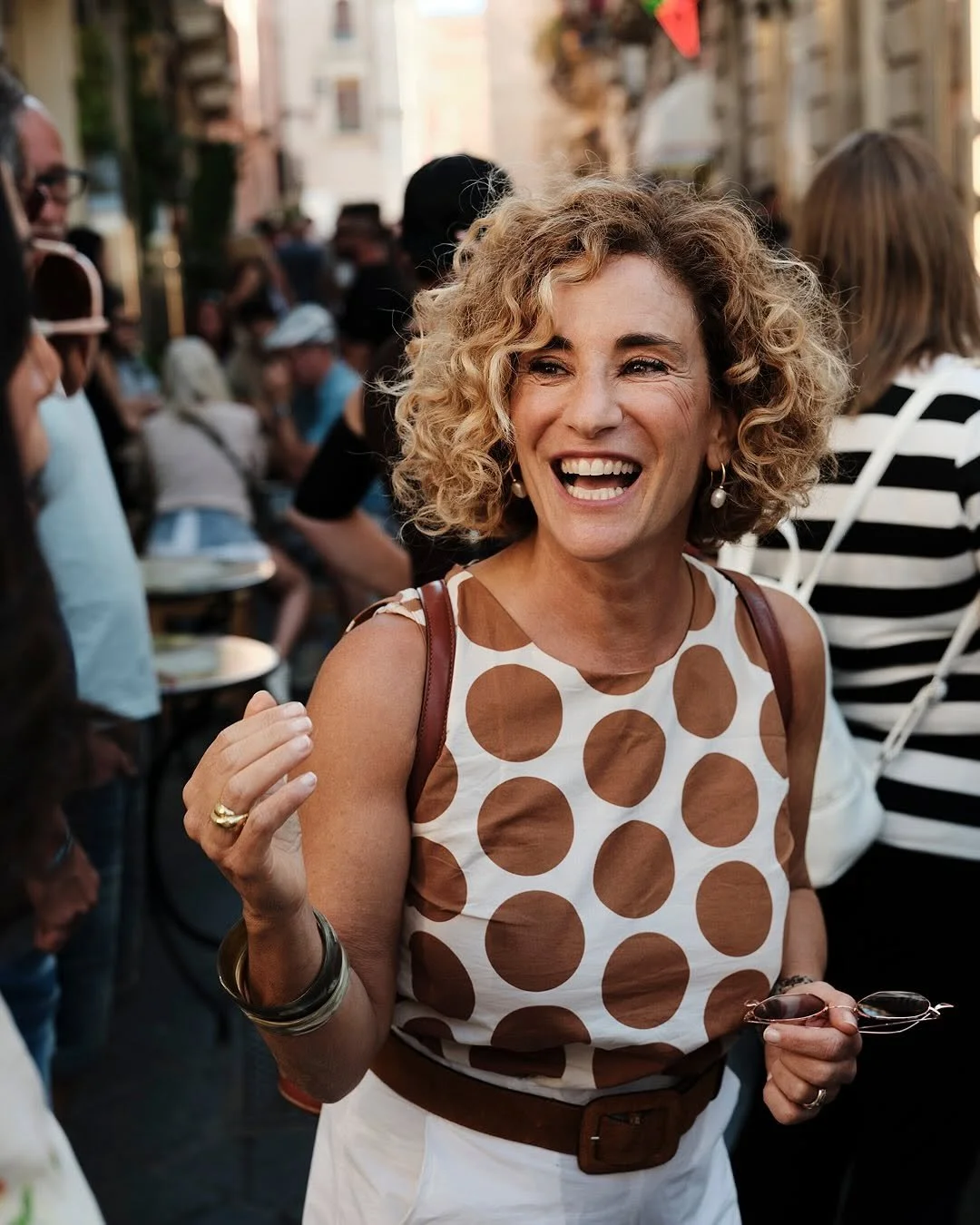 A woman with curly blonde hair smiling and laughing at an outdoor social gathering, holding her glasses in her right hand, wearing a sleeveless top with large brown polka dots and pearl earrings.