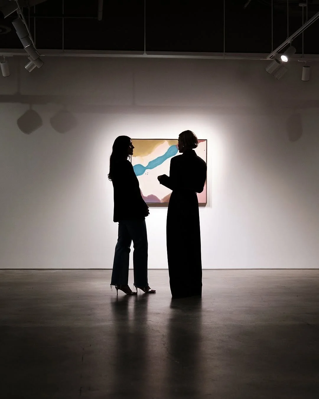 Silhouettes of two people talking in an art gallery with a colorful painting on the wall behind them.