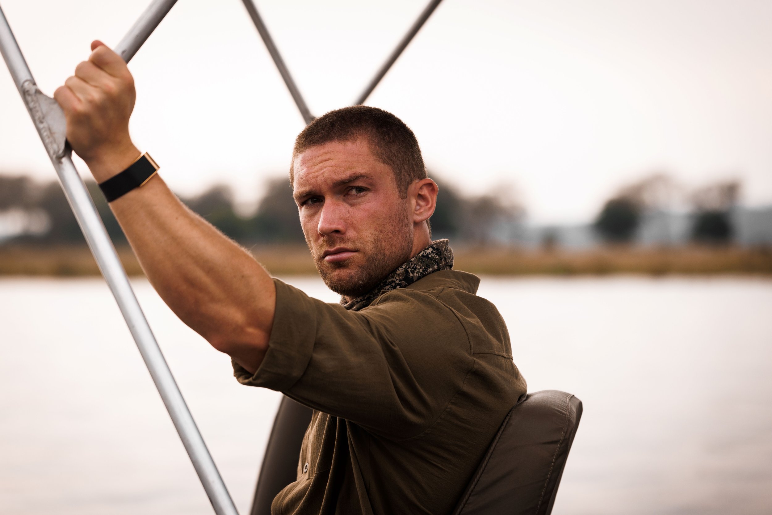 A man sitting in a boat on a lake during sunset, holding onto a metal pole and looking at the camera.