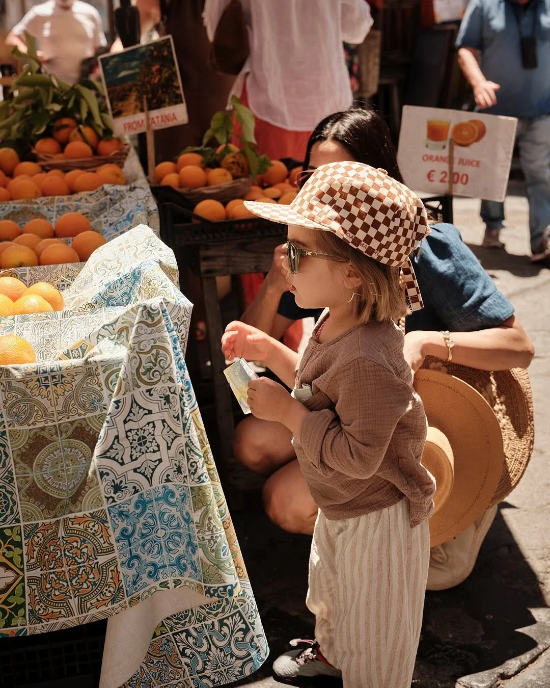 A young girl wearing a checkered sun hat, sunglasses, and a brown shirt shopping for peaches at an outdoor market. There are baskets of peaches and a sign in the background indicating orange juice for 2 euros.