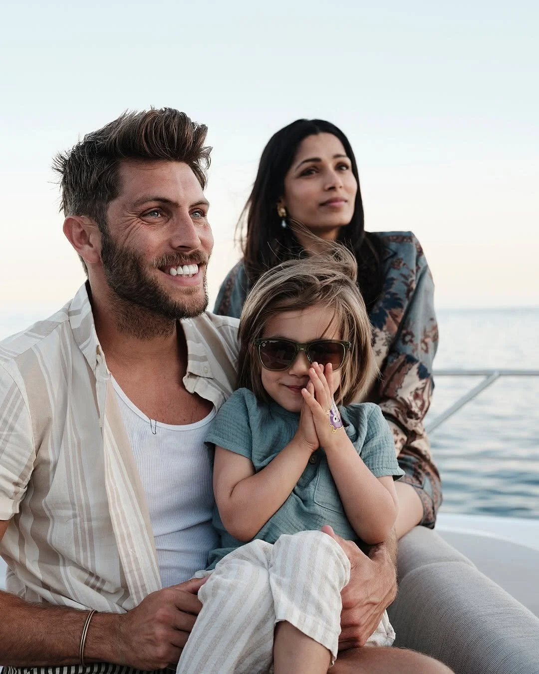 A happy family of three sitting on a boat, with the ocean in the background. The father has brown hair and a beard, wearing a striped shirt. The mother has long black hair and is wearing earrings and a patterned top. The young girl, wearing sunglasses and a romper, is sitting on her dad's lap, with her hands pressed together near her face.
