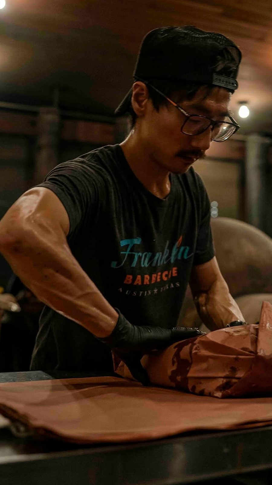 A man wearing a black cap, glasses, and a black t-shirt with 'Franklin Barbecue' logo, preparing meat with gloves in a dimly lit indoor setting.