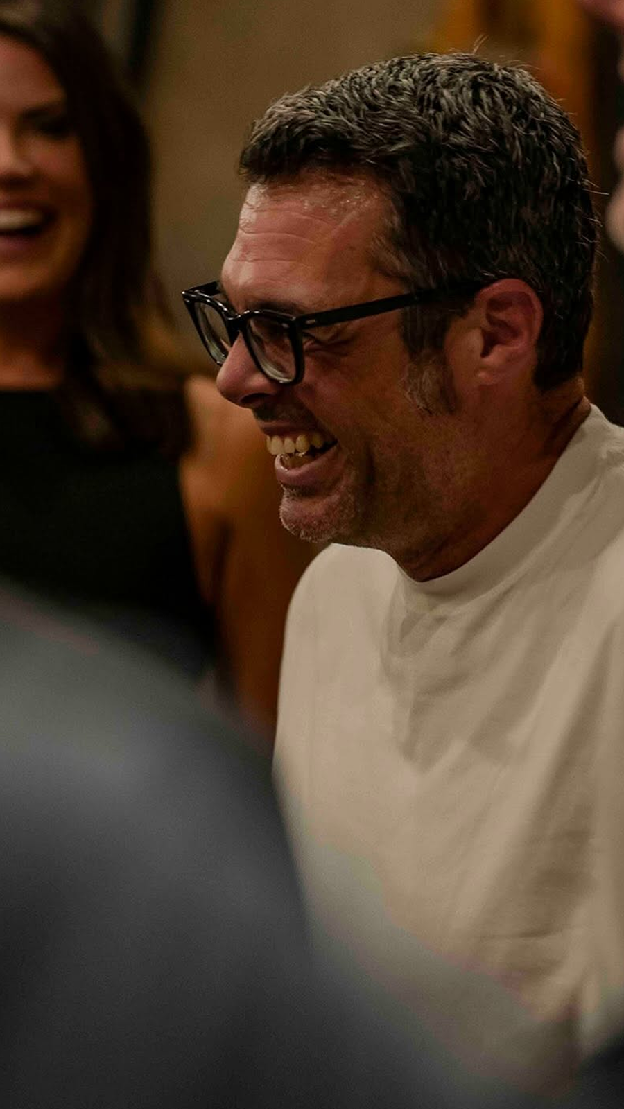 A man with glasses and gray hair, smiling and laughing, wearing a white shirt, in a social setting with people around.