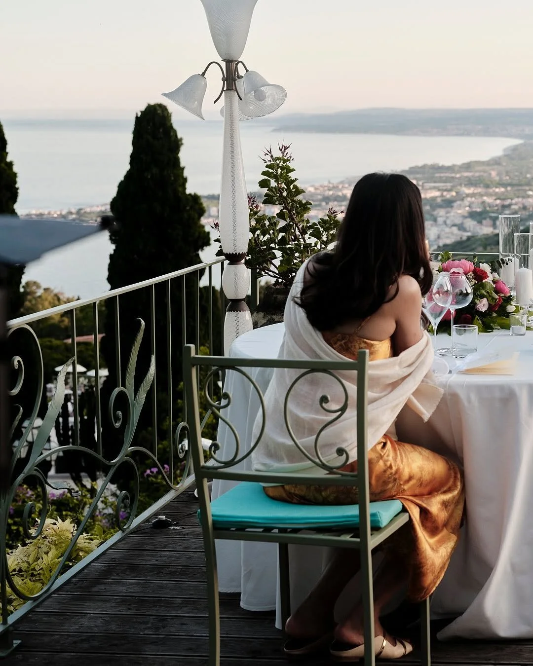 A woman sitting at a dining table on a balcony overlooking a body of water and a distant cityscape, with a floral arrangement and glassware on the table, during sunset.