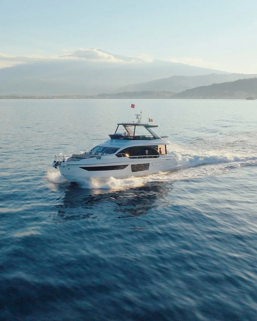 A white luxury yacht sailing across calm water with distant mountains and a partly cloudy sky in the background.