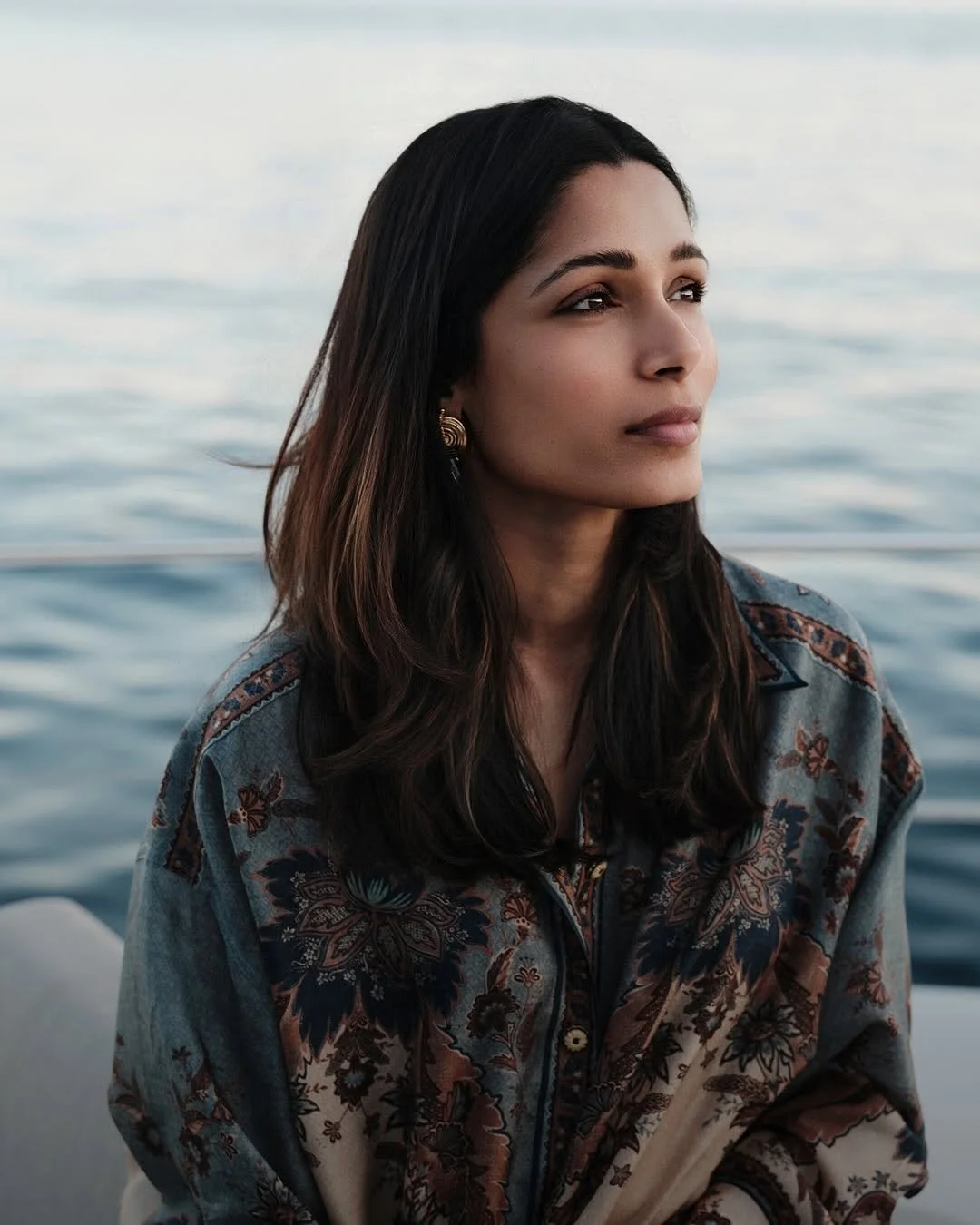 A woman with long dark hair and earrings looking thoughtfully to the side while sitting by the water, wearing a patterned blouse.