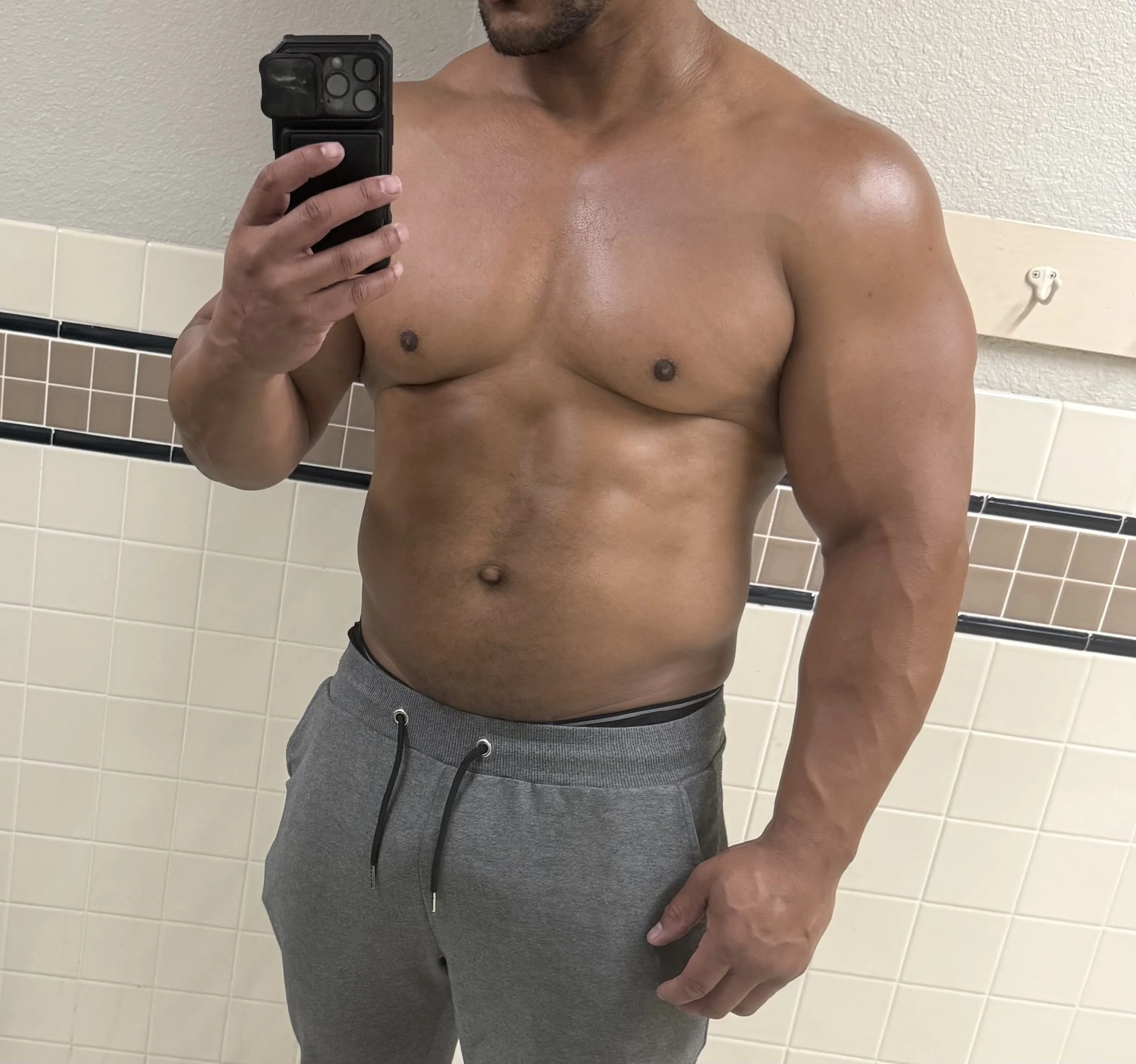 A shirtless man taking a mirror selfie in a bathroom, showing his muscular upper body and wearing gray sweatpants.