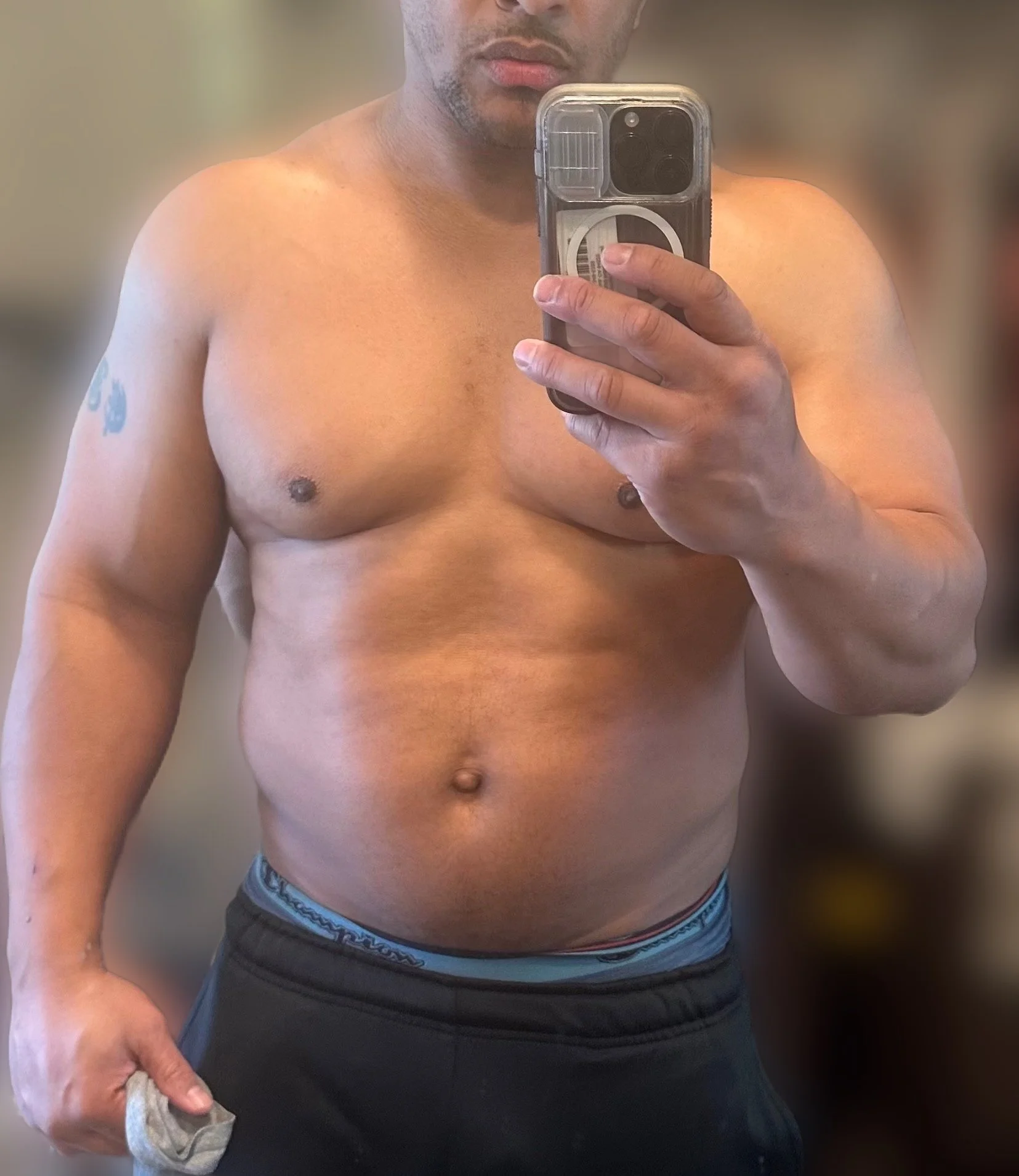 A shirtless person taking a mirror selfie with a smartphone, holding a towel in their left hand, with a blurred background.