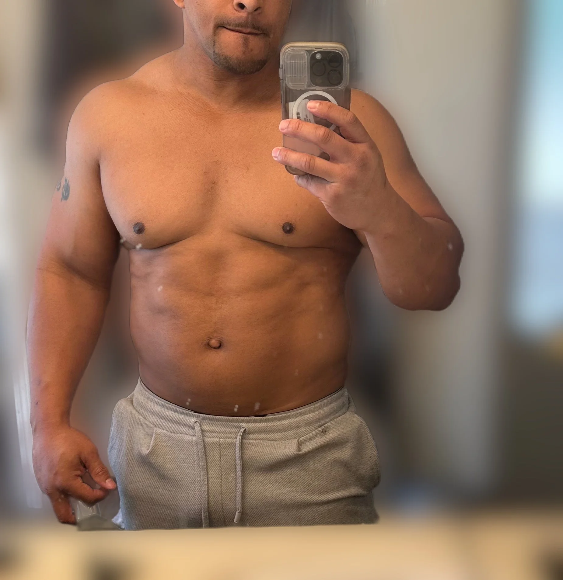 A shirtless man taking a mirror selfie with a smartphone, wearing gray sweatpants, showing his upper body and stomach.