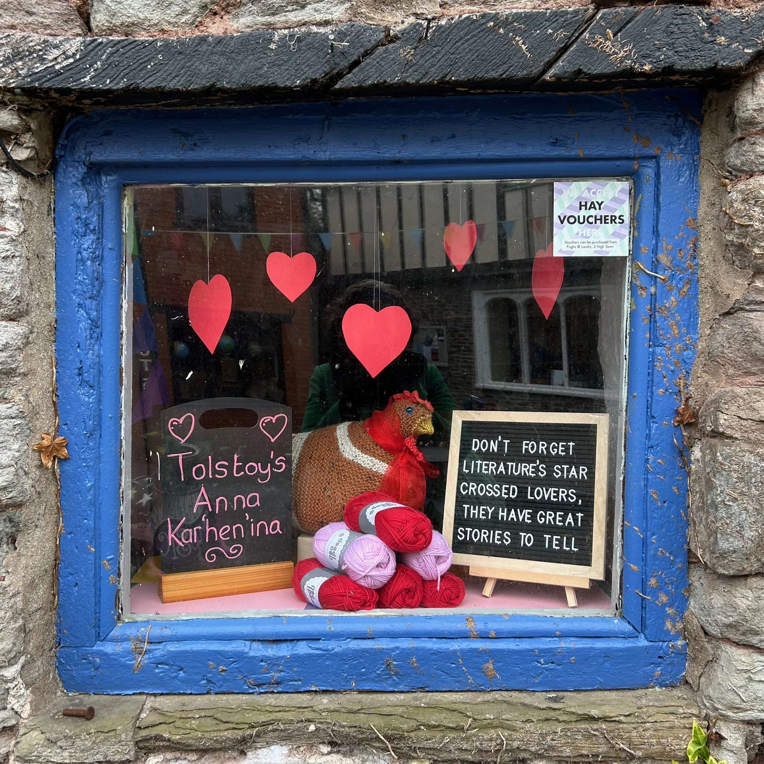 stone building square window with blue trim. Inside the window is a knit hen and piles of red and pink yarn.
