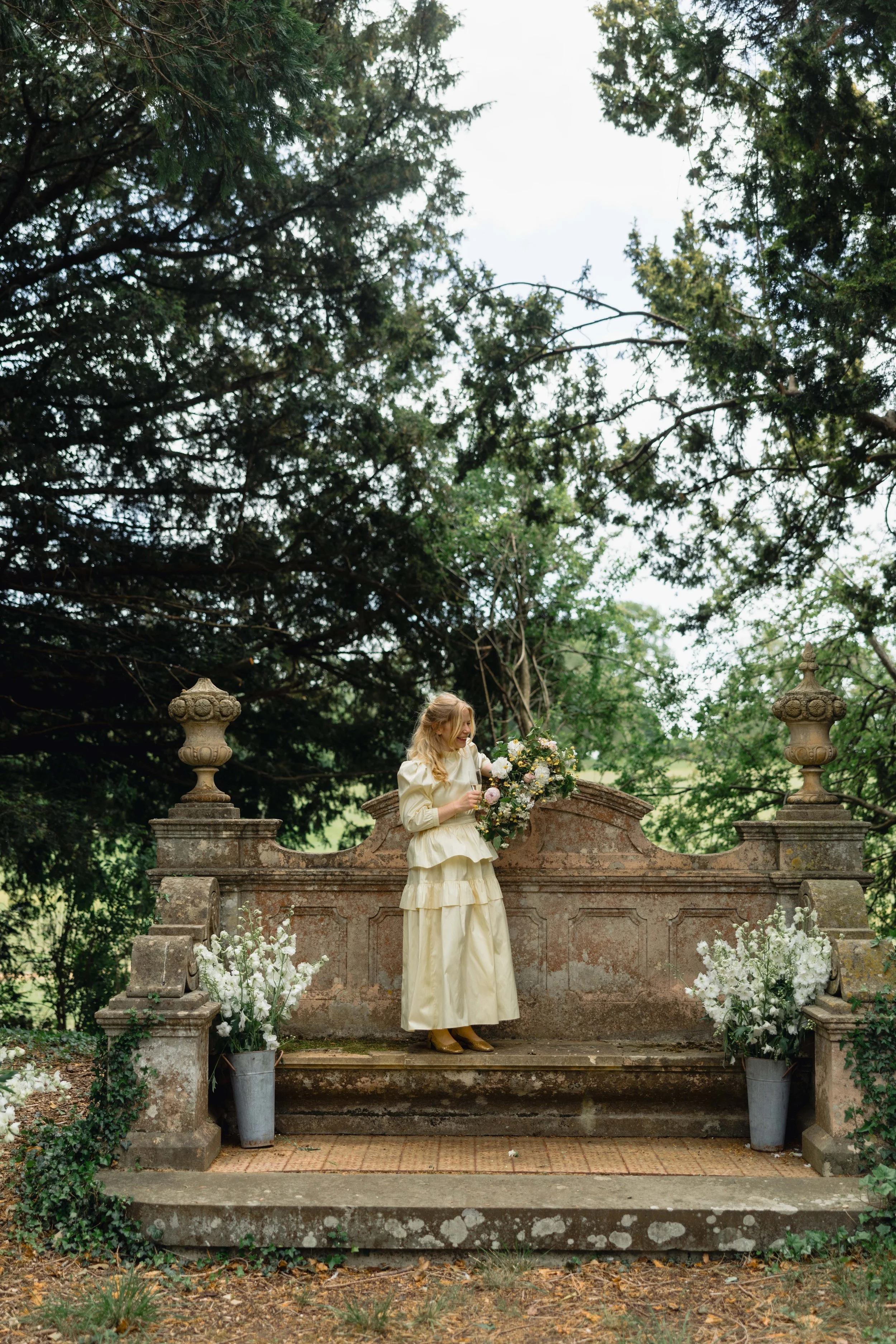A woman in a cream-colored vintage dress holding a bouquet of flowers, standing on an old stone terrace decorated with white flowers and greenery, outdoors among tall trees.