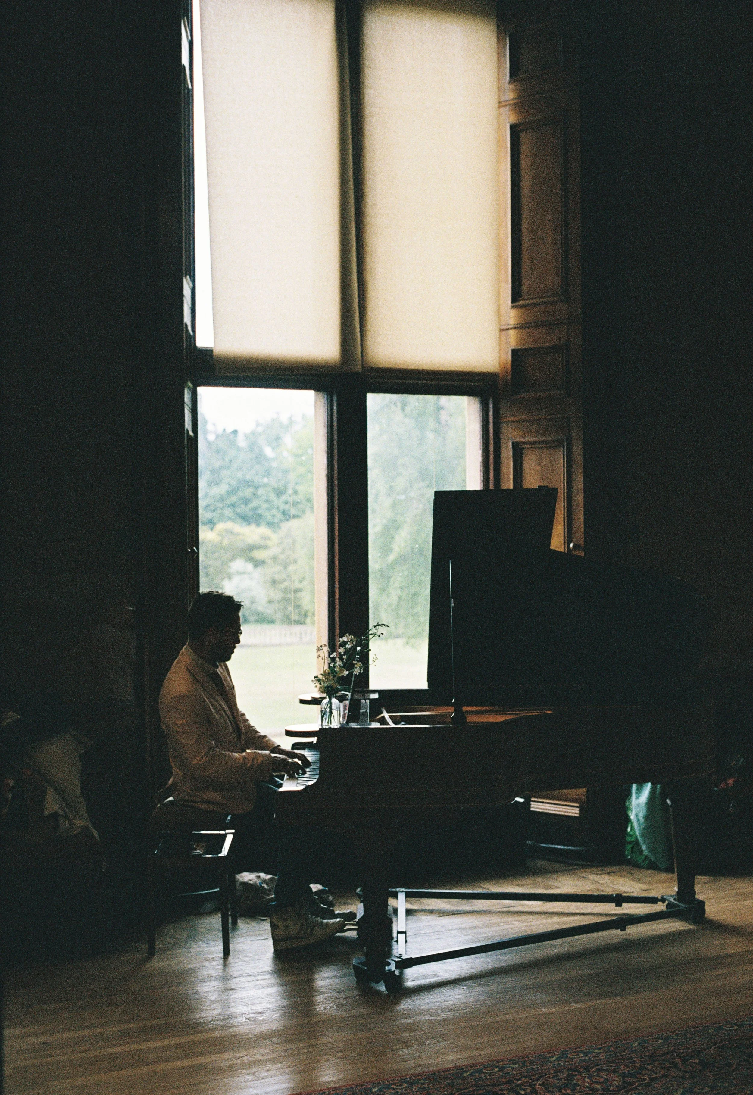 A person playing a grand piano indoors near large windows with beige blinds. The room has wooden walls and bright natural light coming in.