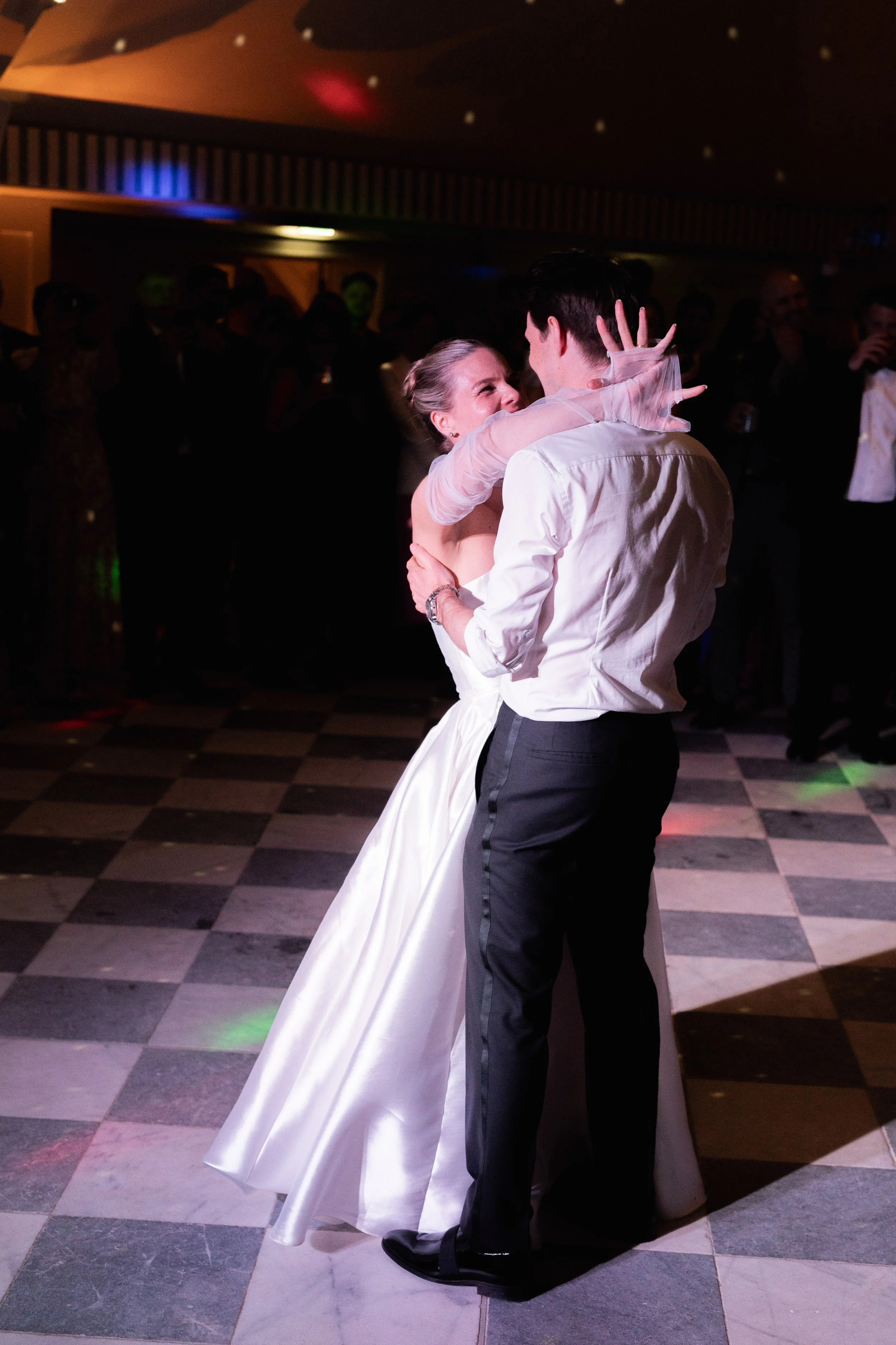 A bride and groom are dancing closely on the wedding dance floor, surrounded by guests in a dimly lit reception hall.