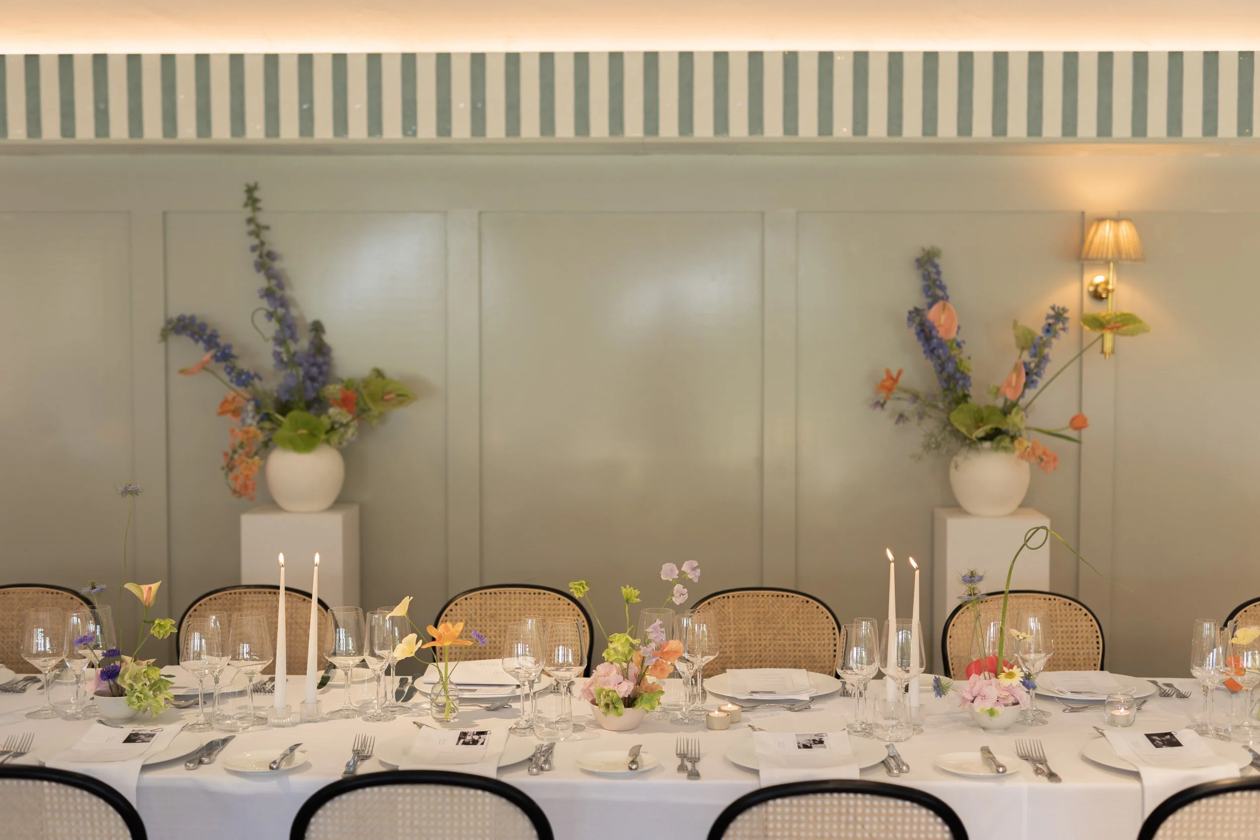 A decorated dining table set for a formal event with floral centerpieces, candles, glassware, and cutlery, in a room with light-colored walls, large vases with purple and pink flowers, and a wall sconce with a small lampshade.