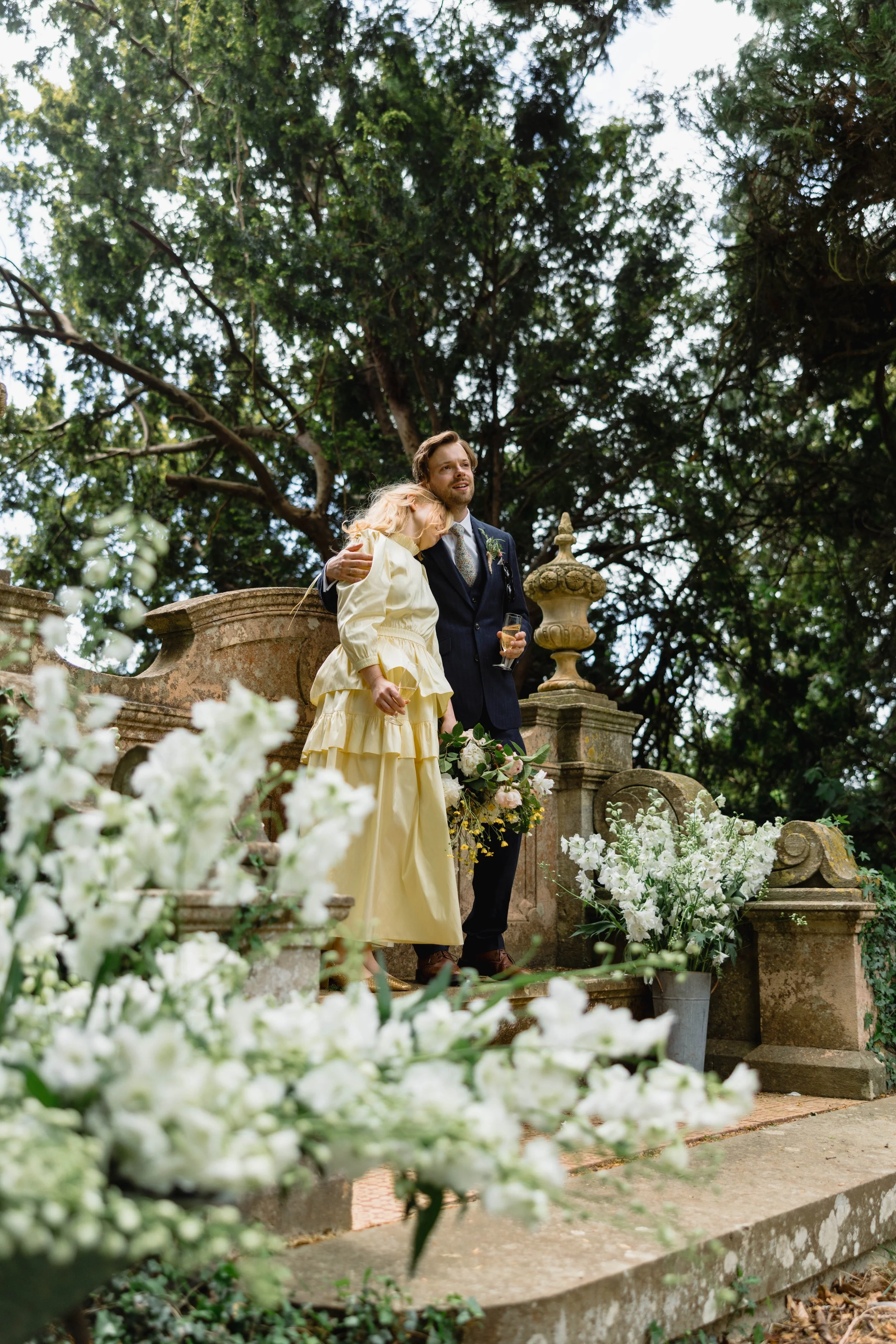 A couple at a wedding or formal event, standing on an ornate stone platform surrounded by white flowers and greenery, with large trees in the background. The woman is in a yellow dress, leaning on the man's shoulder, who is in a dark suit holding a g