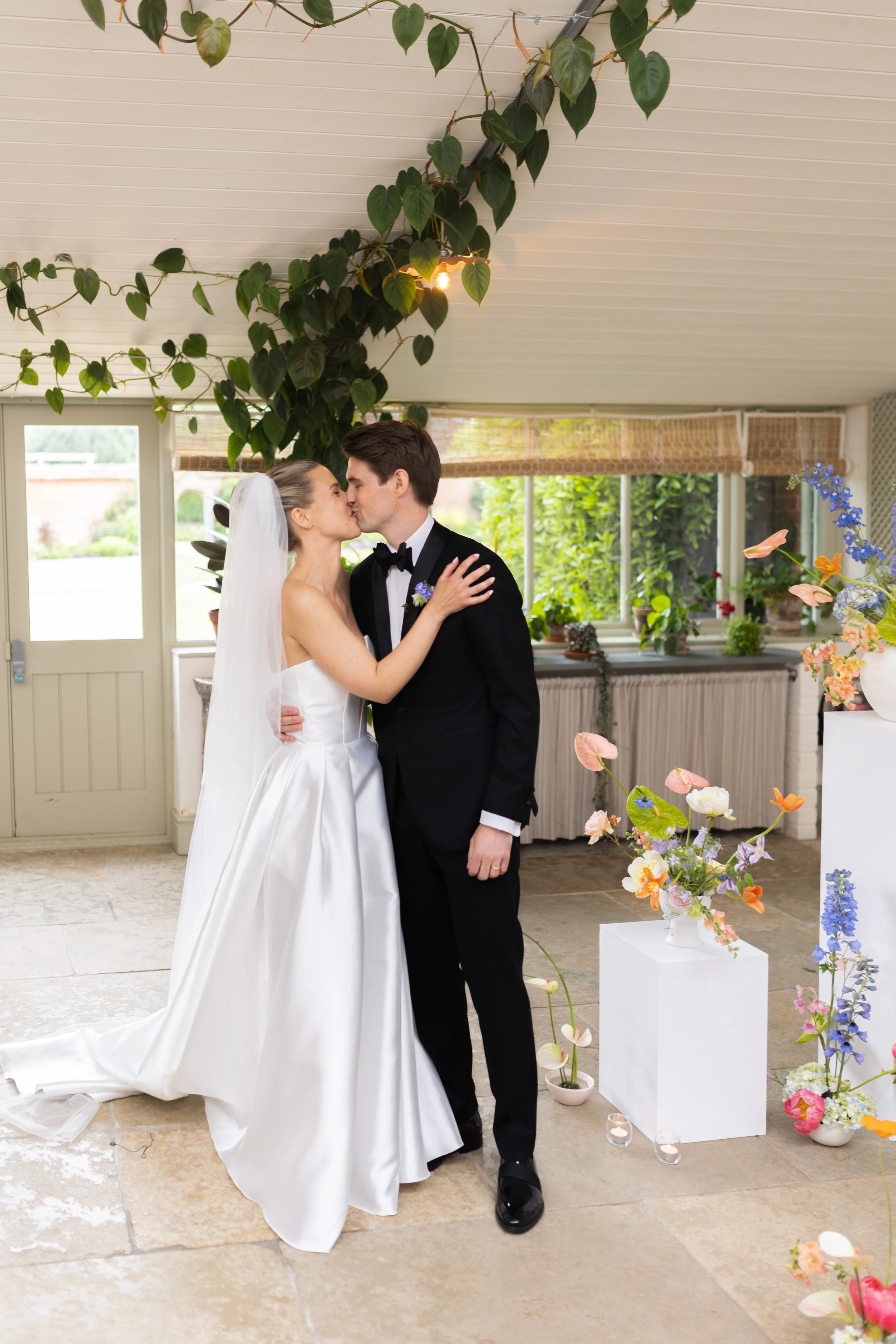 A bride and groom kissing during their wedding, surrounded by flowers and greenery inside a decorated venue.
