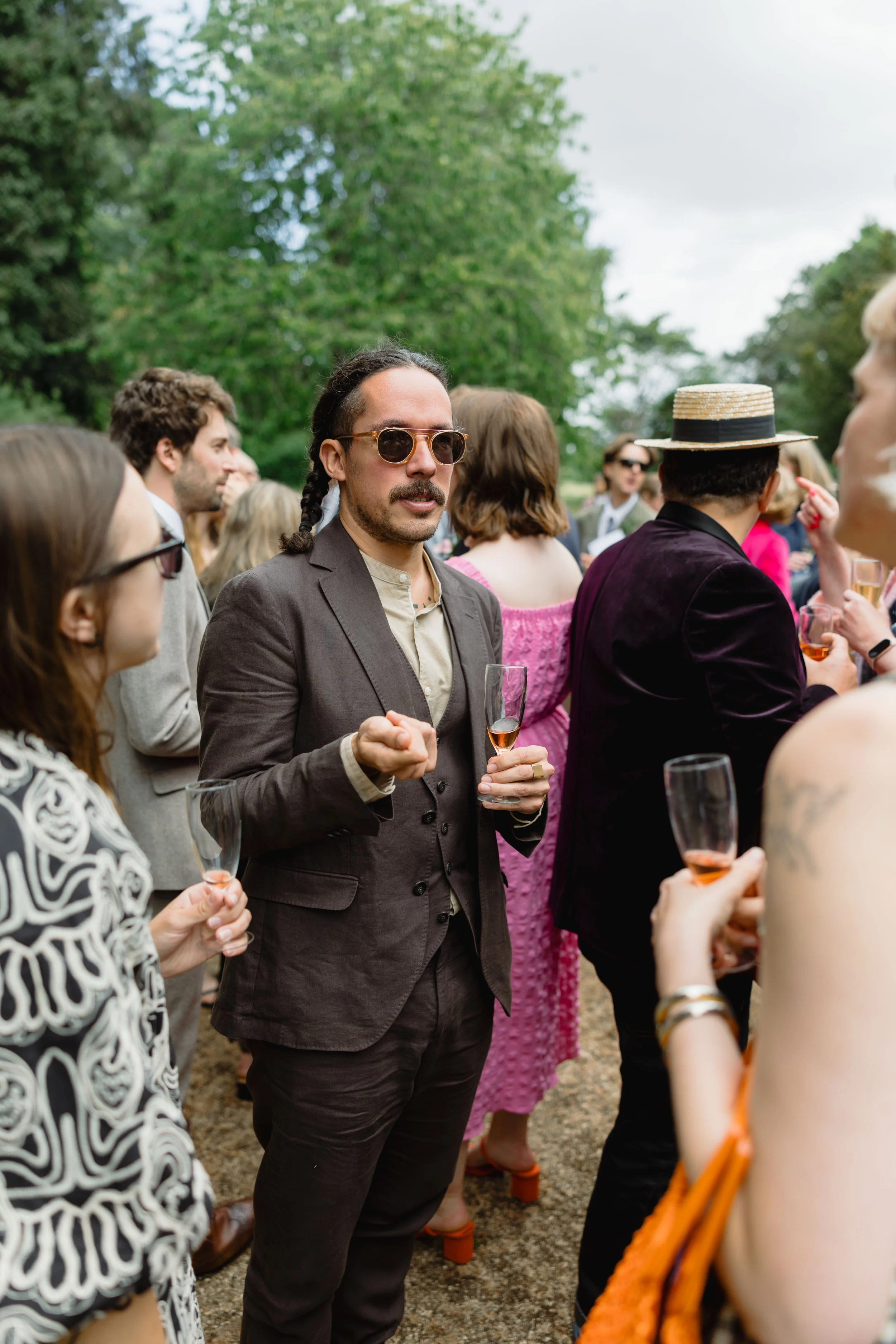 Group of people at an outdoor gathering, some holding drinks, with lush green trees in the background.
