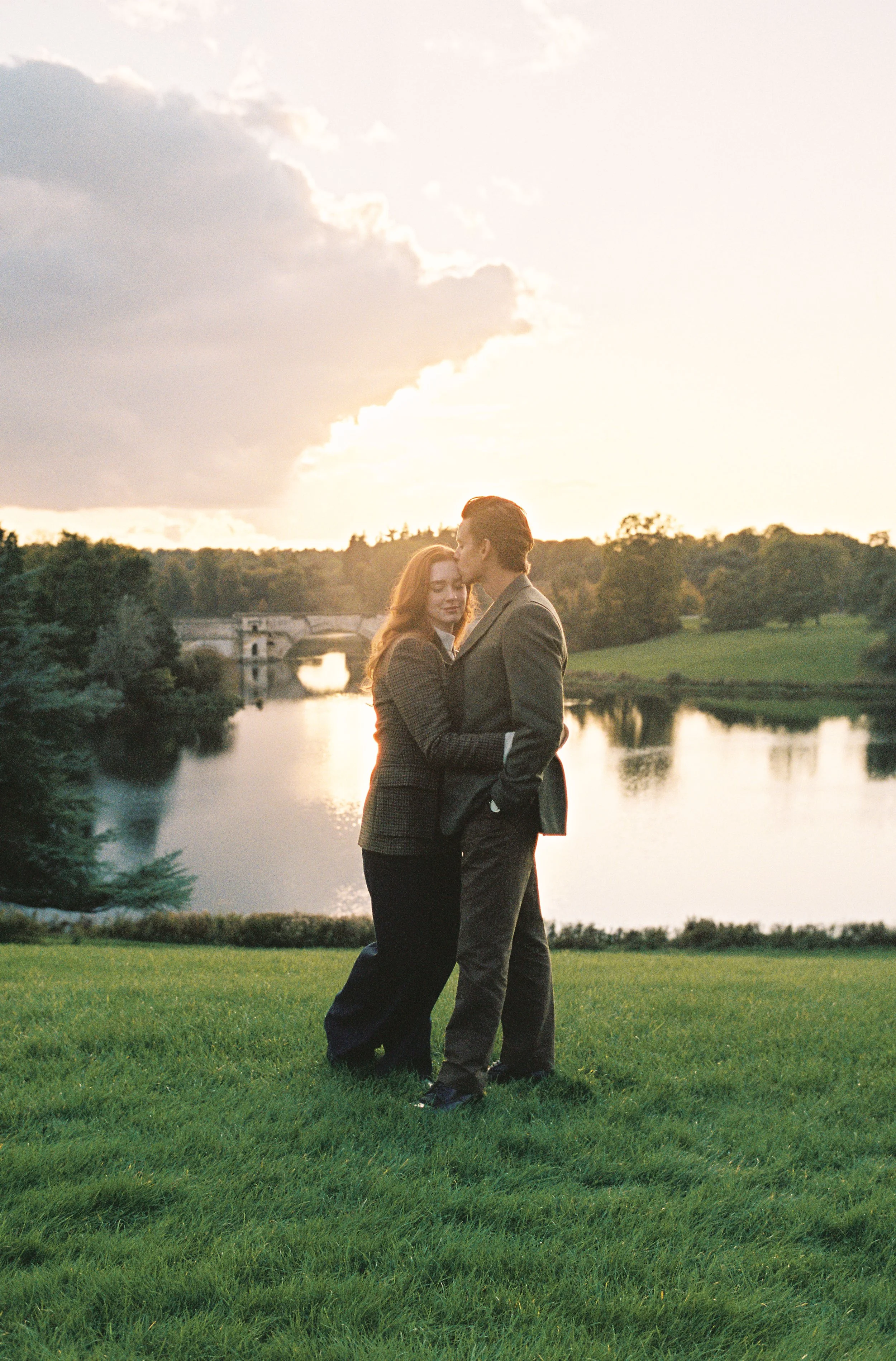 A couple dressed in formal attire embracing and sharing a kiss by a river at sunset, with a bridge in the background and lush green landscape.