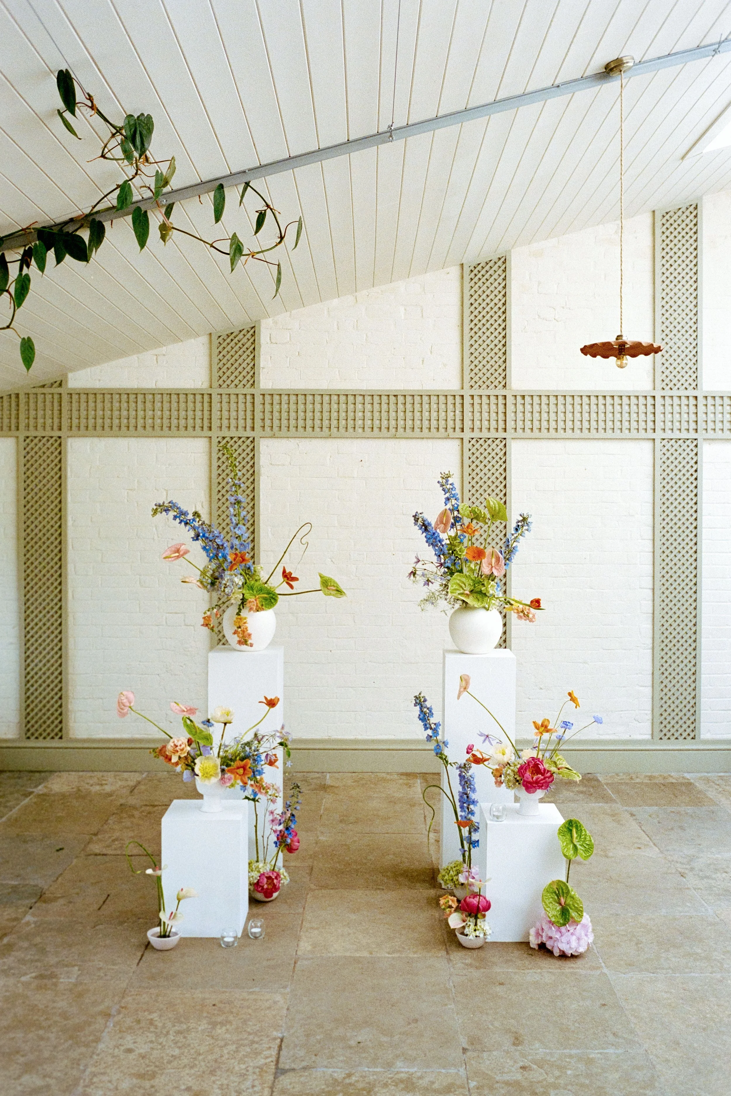 Indoor display of four white vases containing colorful flowers on white pedestals, set against a white brick wall with decorative wooden lattice panels.