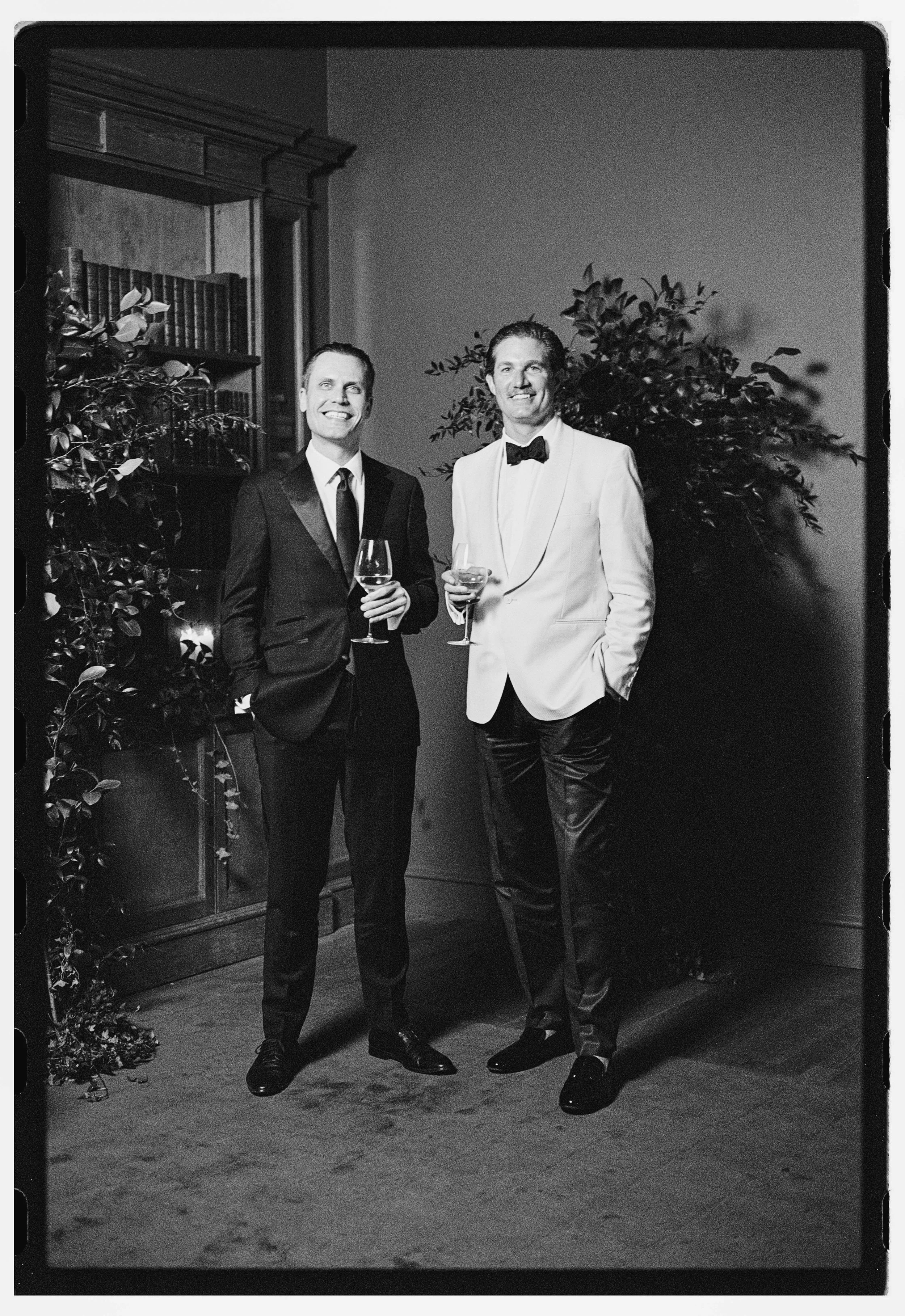Two men in tuxedos standing indoors with wine glasses, smiling, with a bookshelf and potted plants in the background.