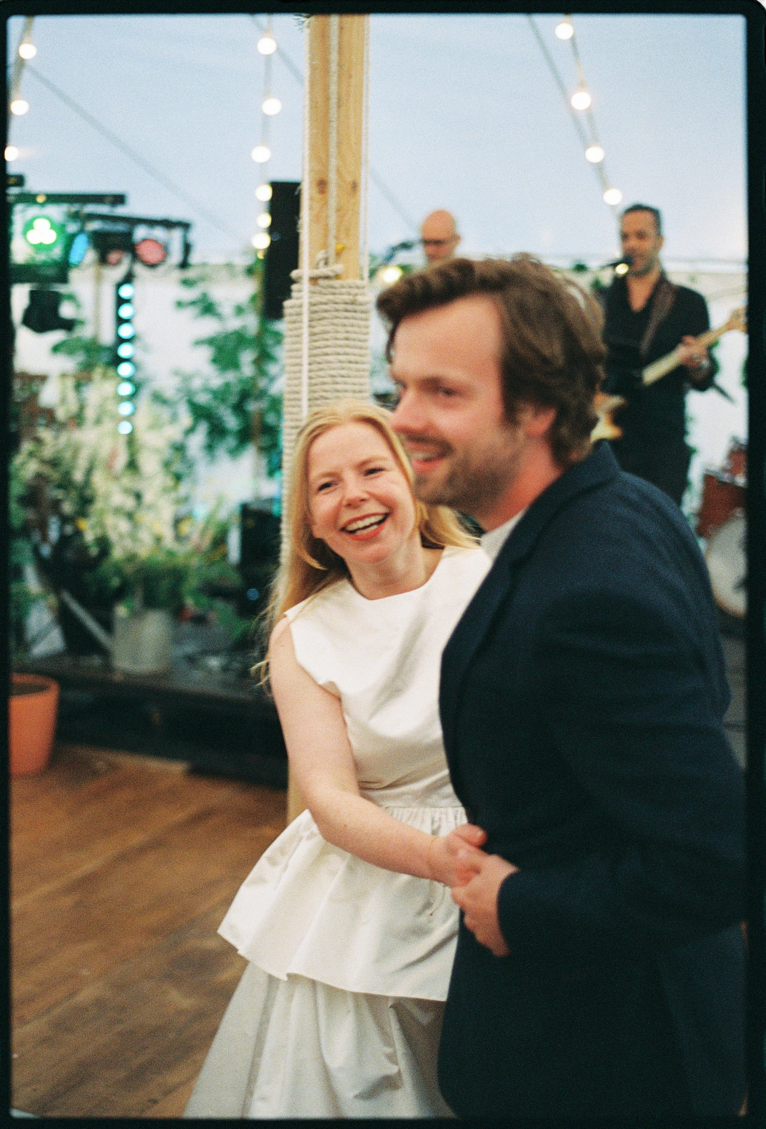 A smiling woman in a white dress dancing with a man in a dark suit at an outdoor event, with a band playing in the background.