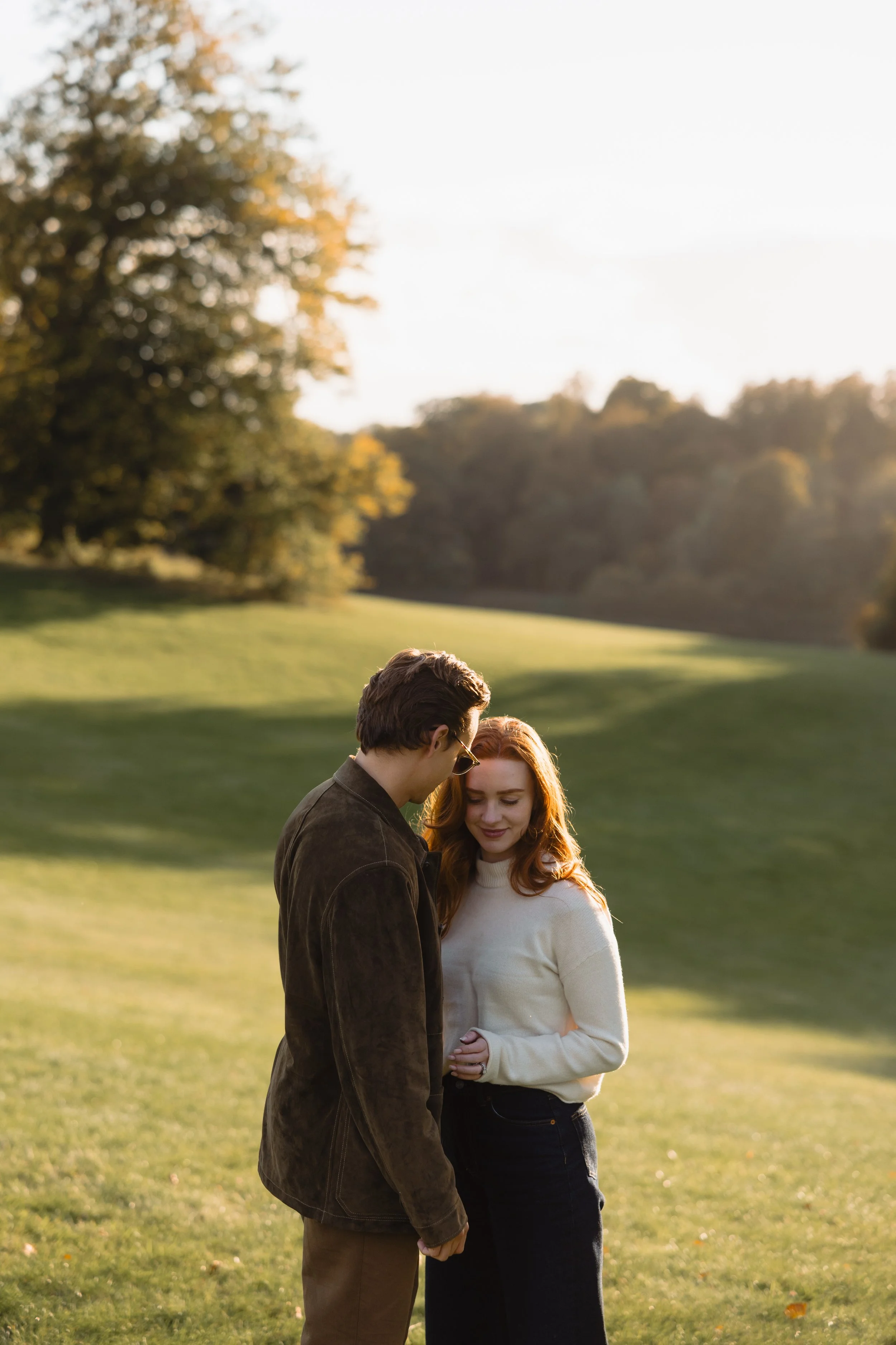A man and woman standing close together in a grassy park, smiling and sharing a moment during sunset.