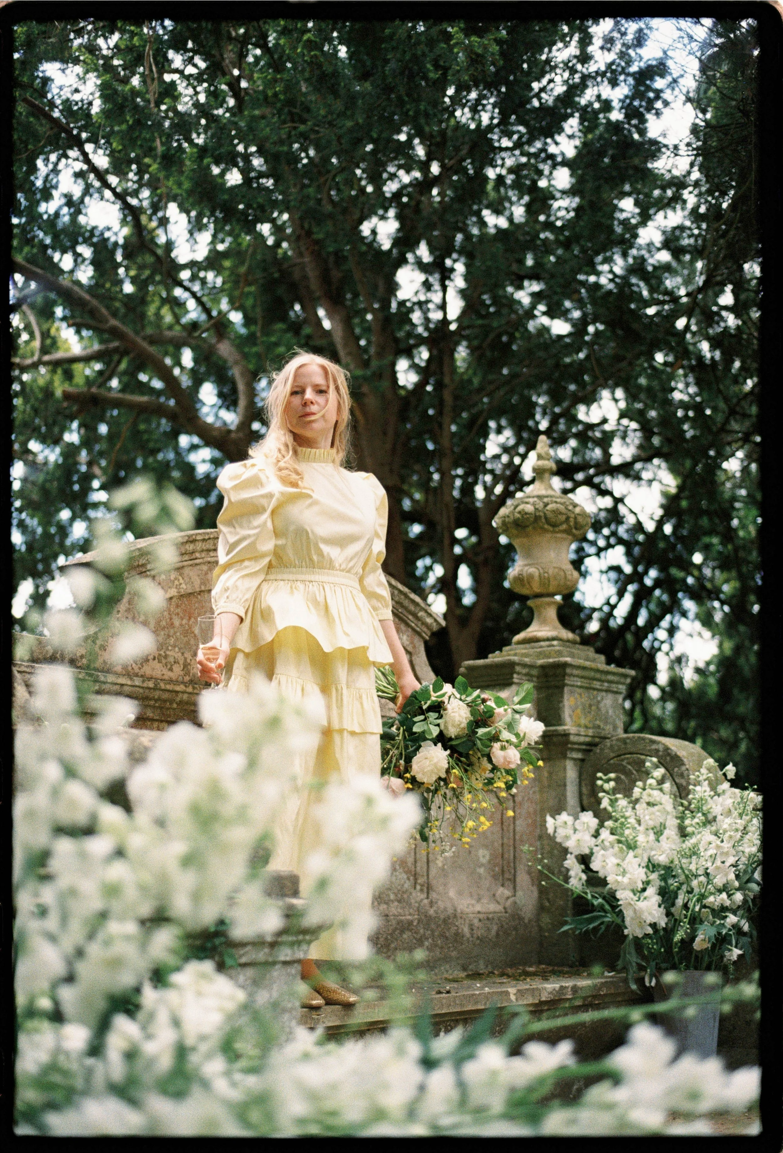 Woman in yellow dress holding a bouquet, standing outdoors on a stone staircase surrounded by white flowers, with large trees in the background.