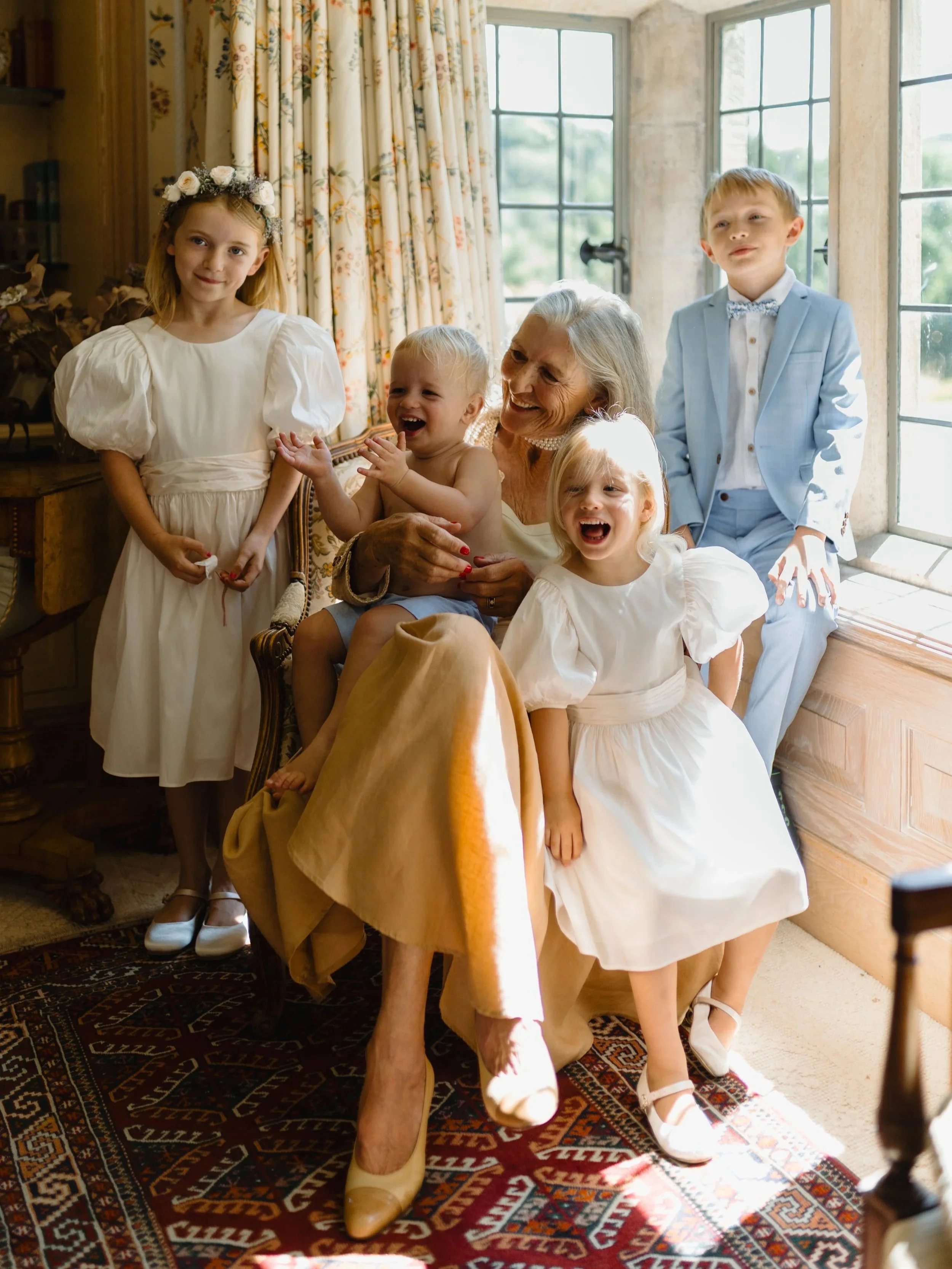 A smiling elderly woman with gray hair surrounded by children in white dresses and a boy in a light blue suit, sitting by a large window in a cozy room.