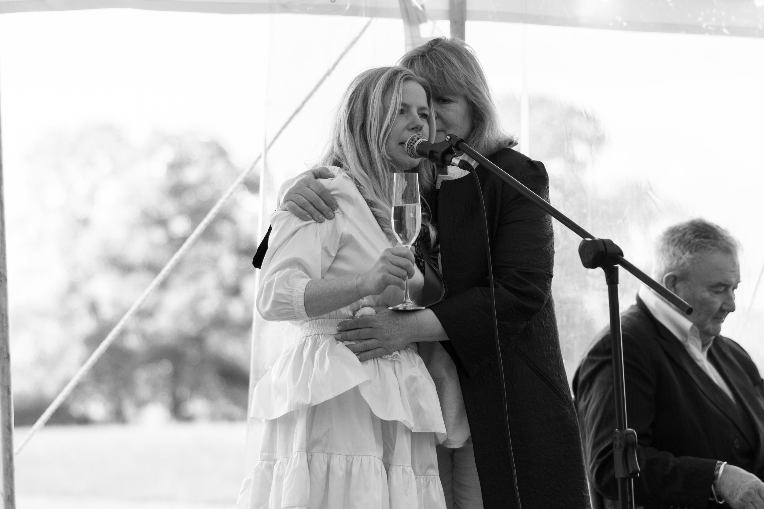 Two women embrace while one holds a champagne glass, singing or speaking into a microphone during a celebration or event, with a man seated in the background.
