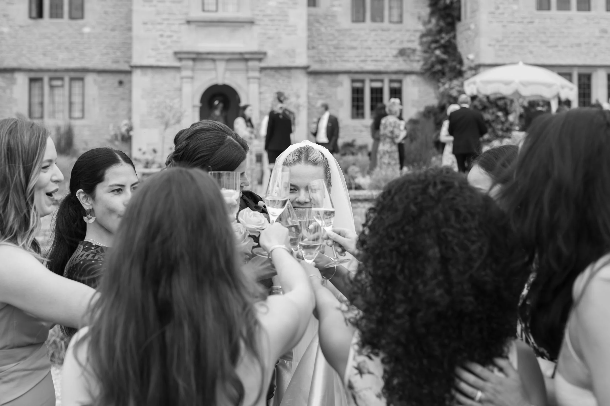 A group of women toasting with champagne glasses at a wedding reception outdoors, with a bride wearing a veil in the center.