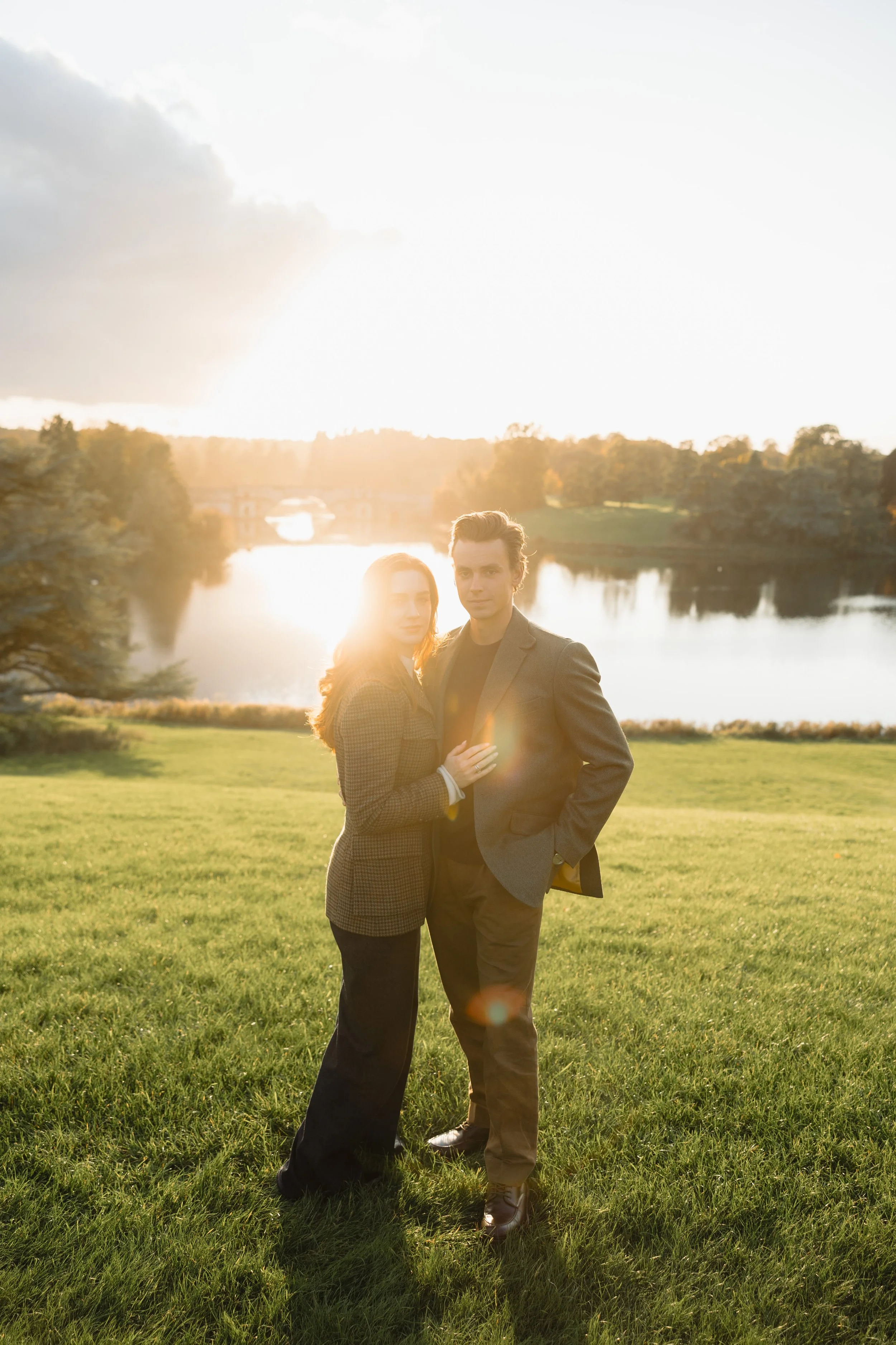 A man and woman standing close together outdoors during sunset by a lake, with trees and a bridge in the background.