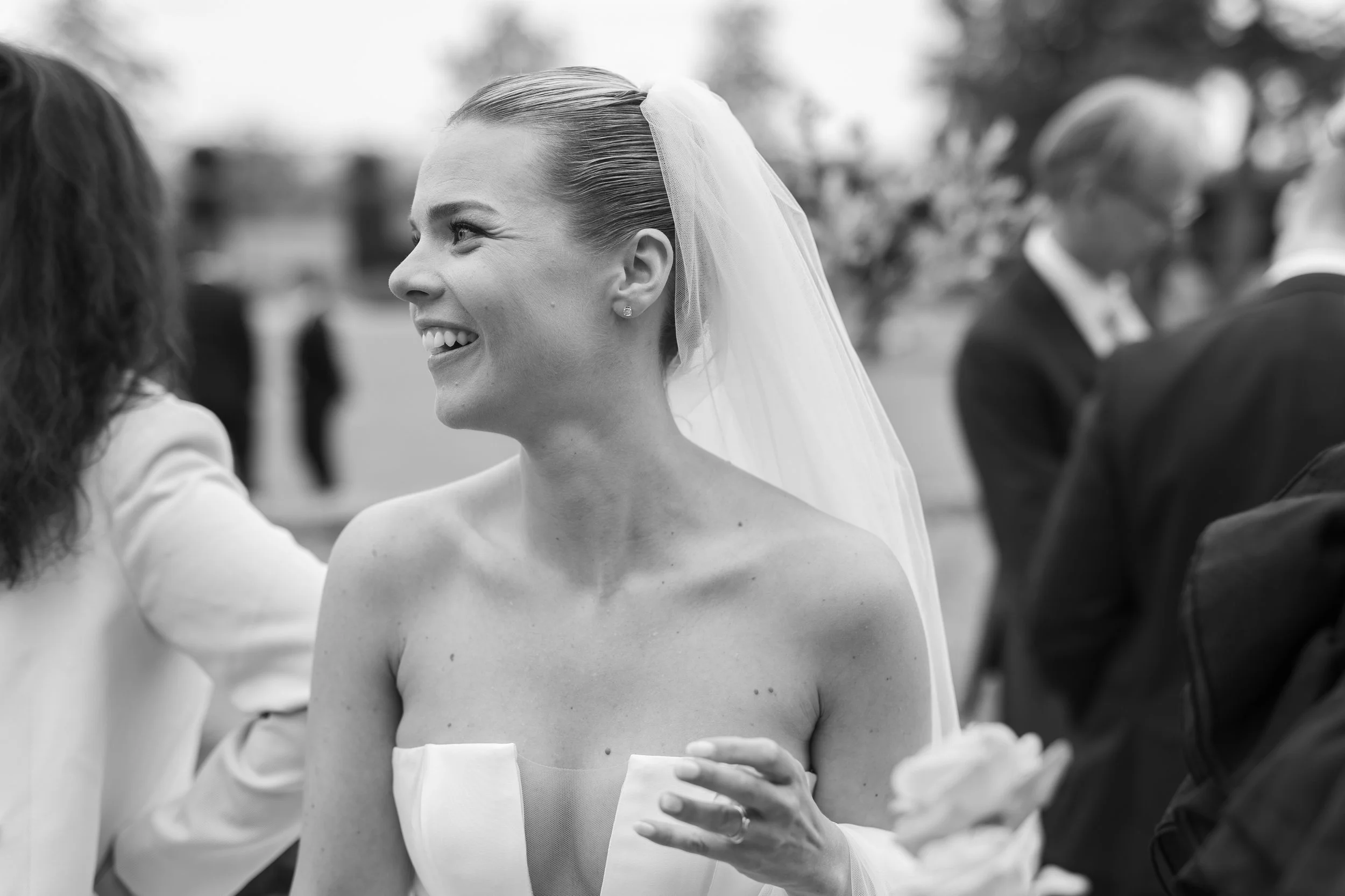 A smiling bride wearing a strapless wedding gown and veil, standing outside at a wedding reception, engaged in conversation with someone off-camera.