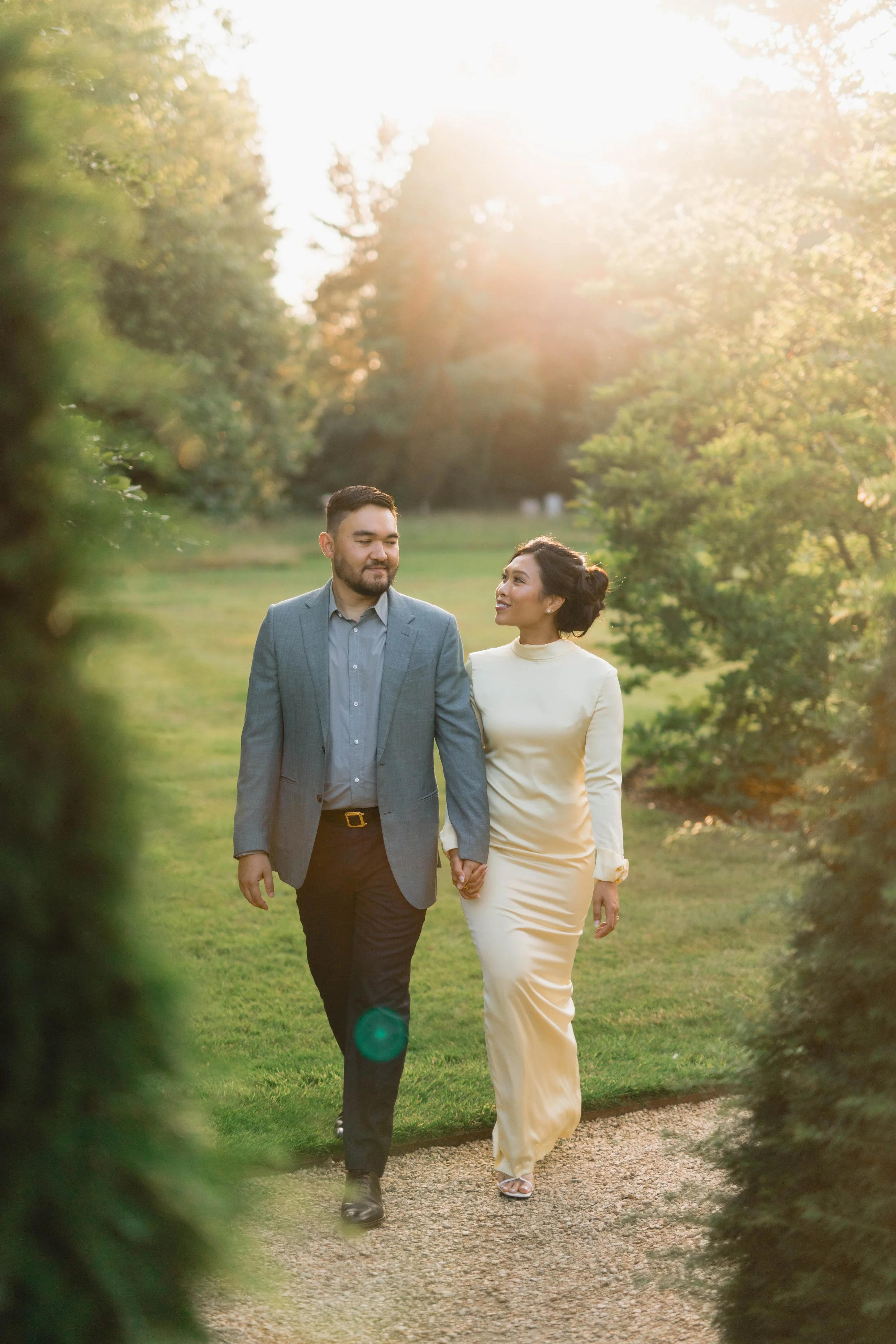 A couple holding hands and walking along a gravel path through a park or garden during sunset, with trees and lush greenery on either side and sunlight filtering through the leaves.
