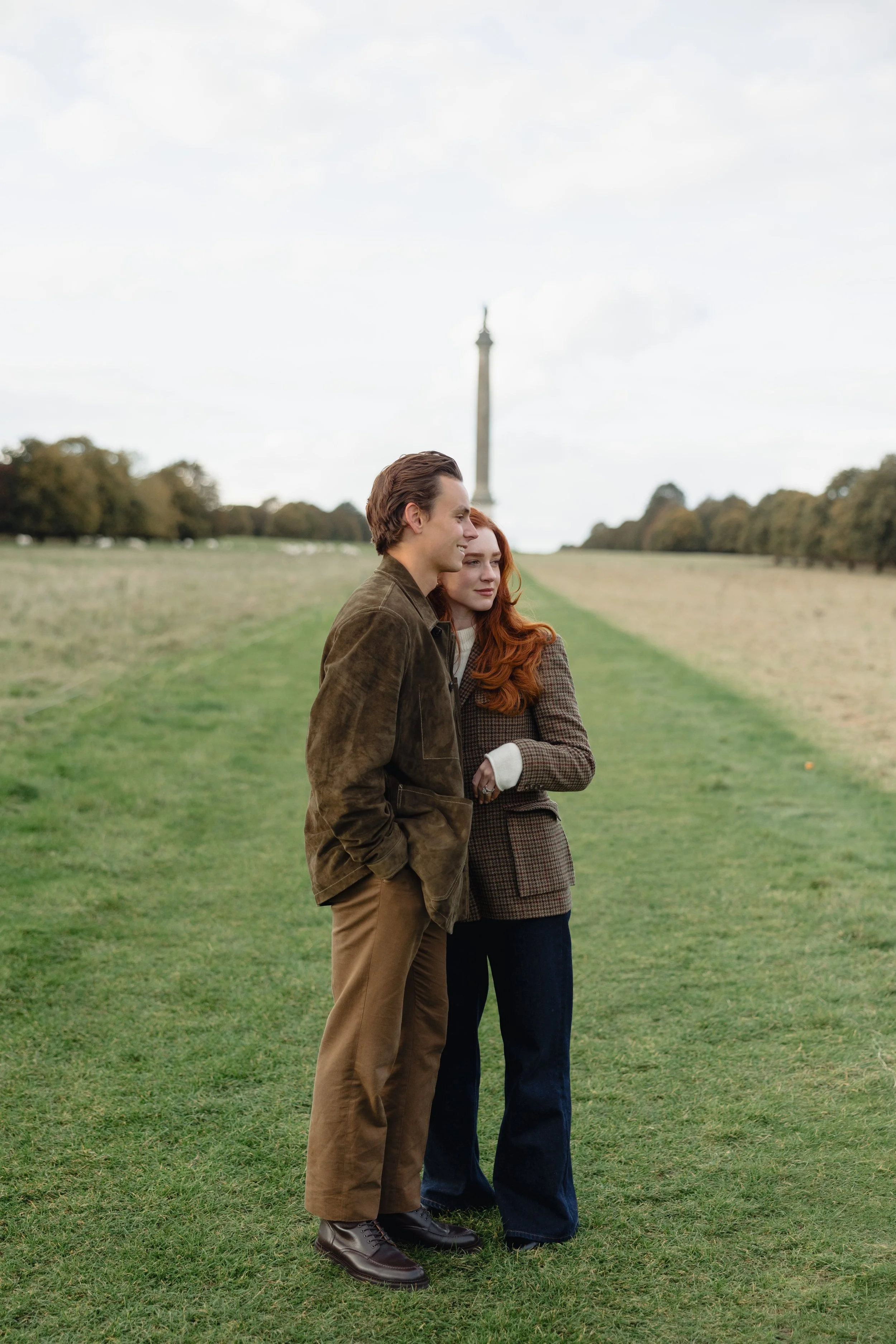A young man and woman standing together on a grassy field with a column monument in the background.