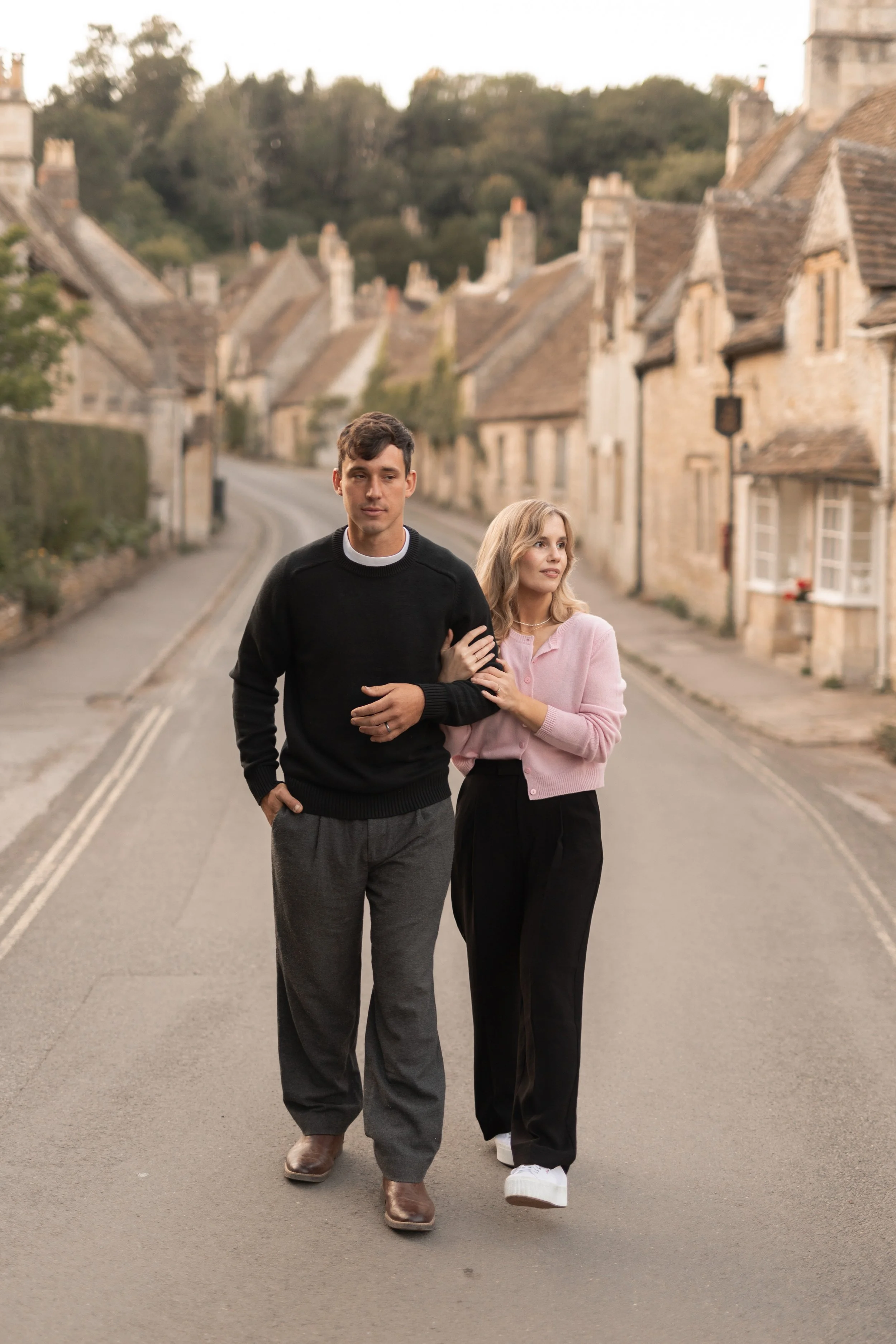 A young man and woman walking arm-in-arm down a quiet, cobblestone street in a picturesque village with stone cottages. The man is dressed in a black sweater and gray trousers, while the woman wears a pink cardigan and black pants. The scene is set d