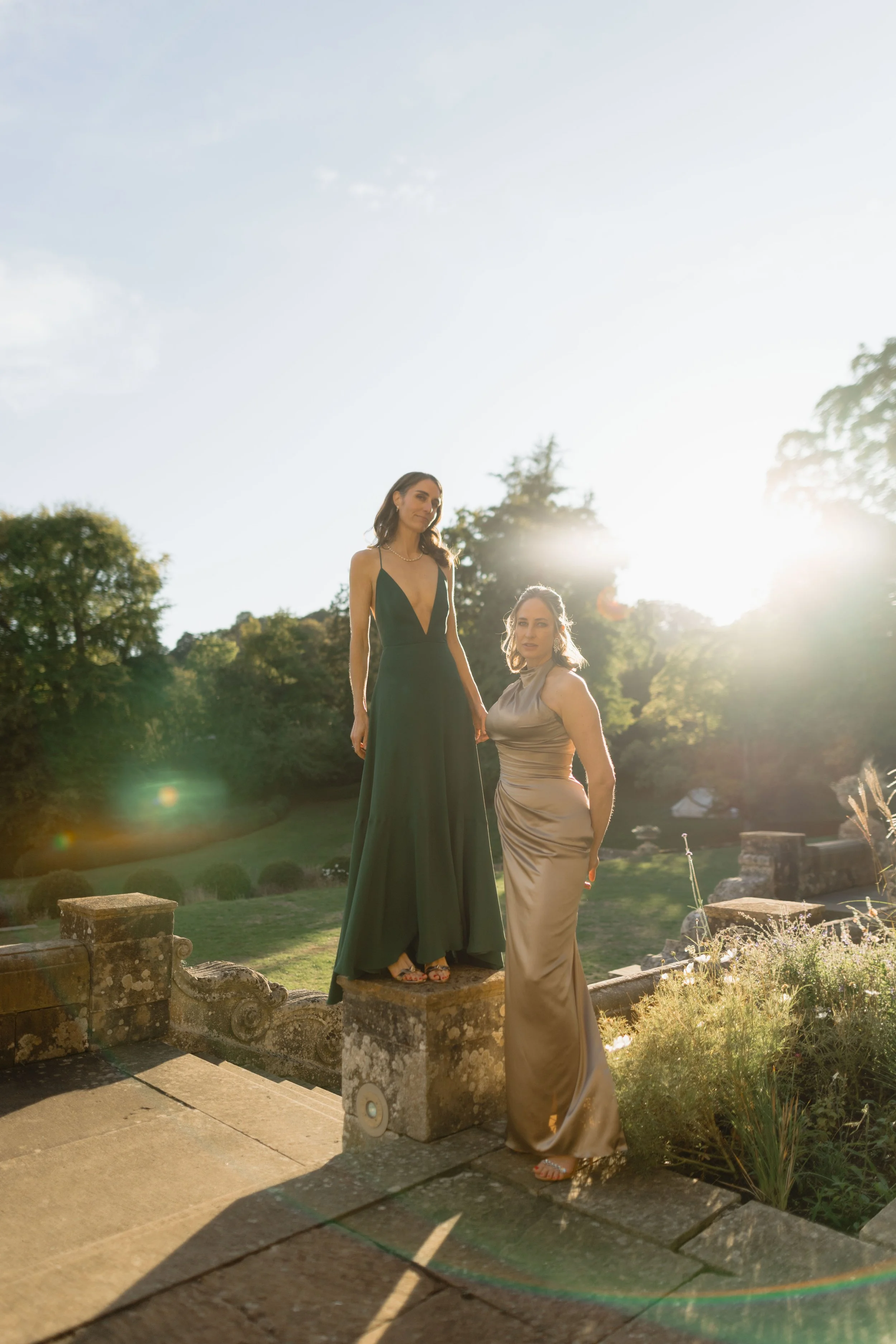 Two women in formal dresses posing outdoors during sunset, with one standing on a stone ledge and the other on the ground, in a garden setting.