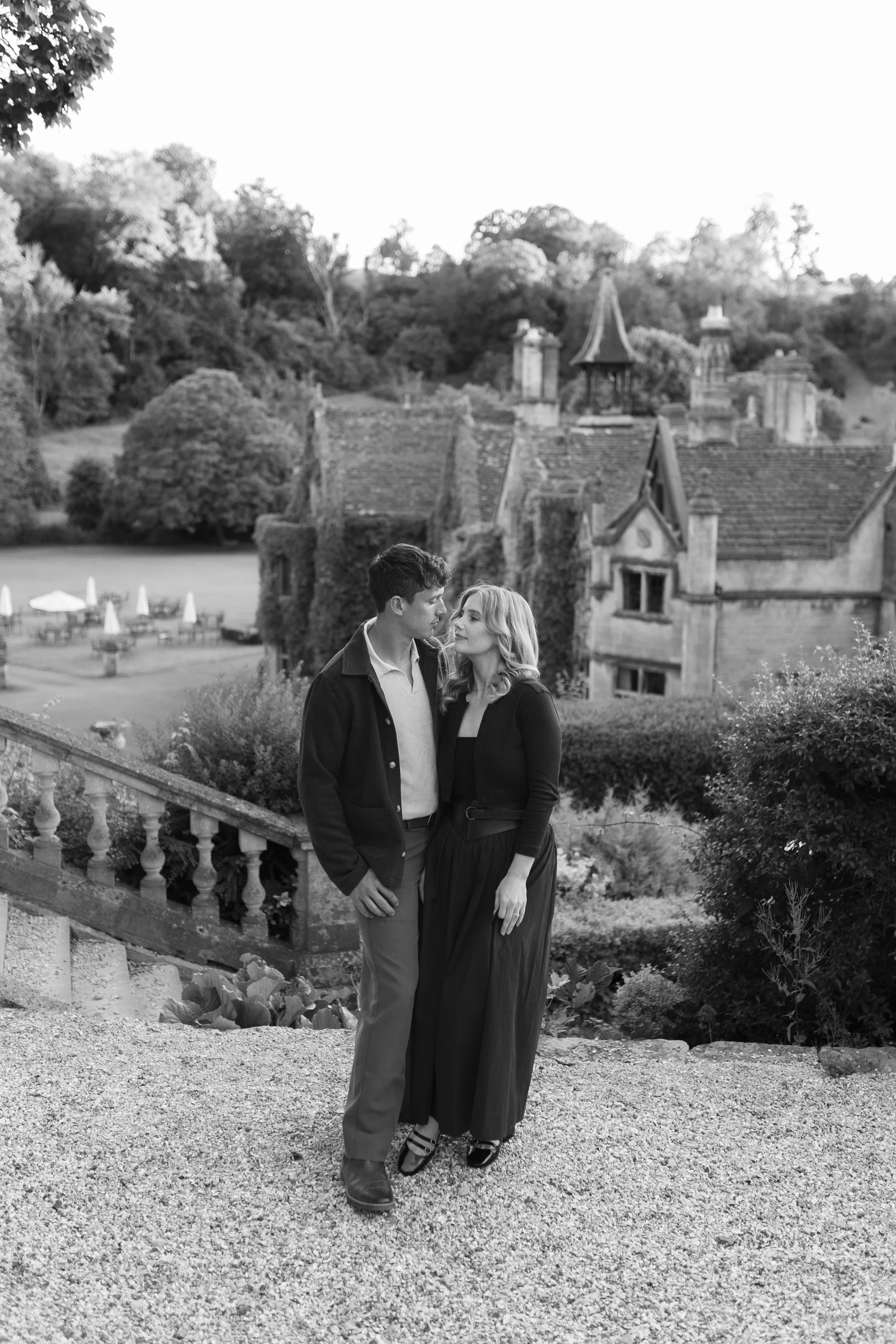A black-and-white photo of a young couple standing close on a gravel path in front of a castle-like building with lush greenery and trees in the background.