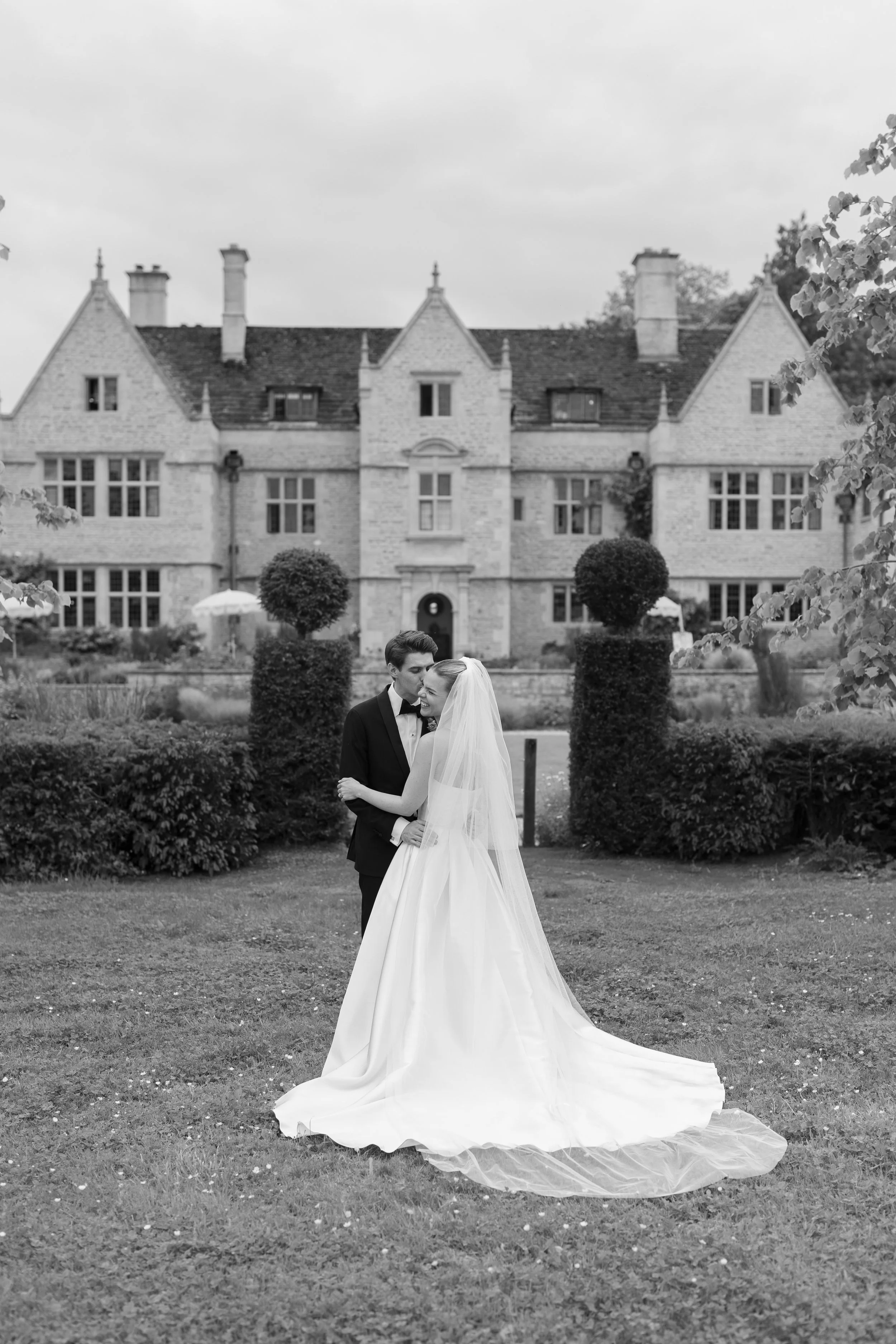 Black and white photograph of a bride and groom in wedding attire embracing on a lawn in front of a large, historic mansion with manicured bushes and trees.