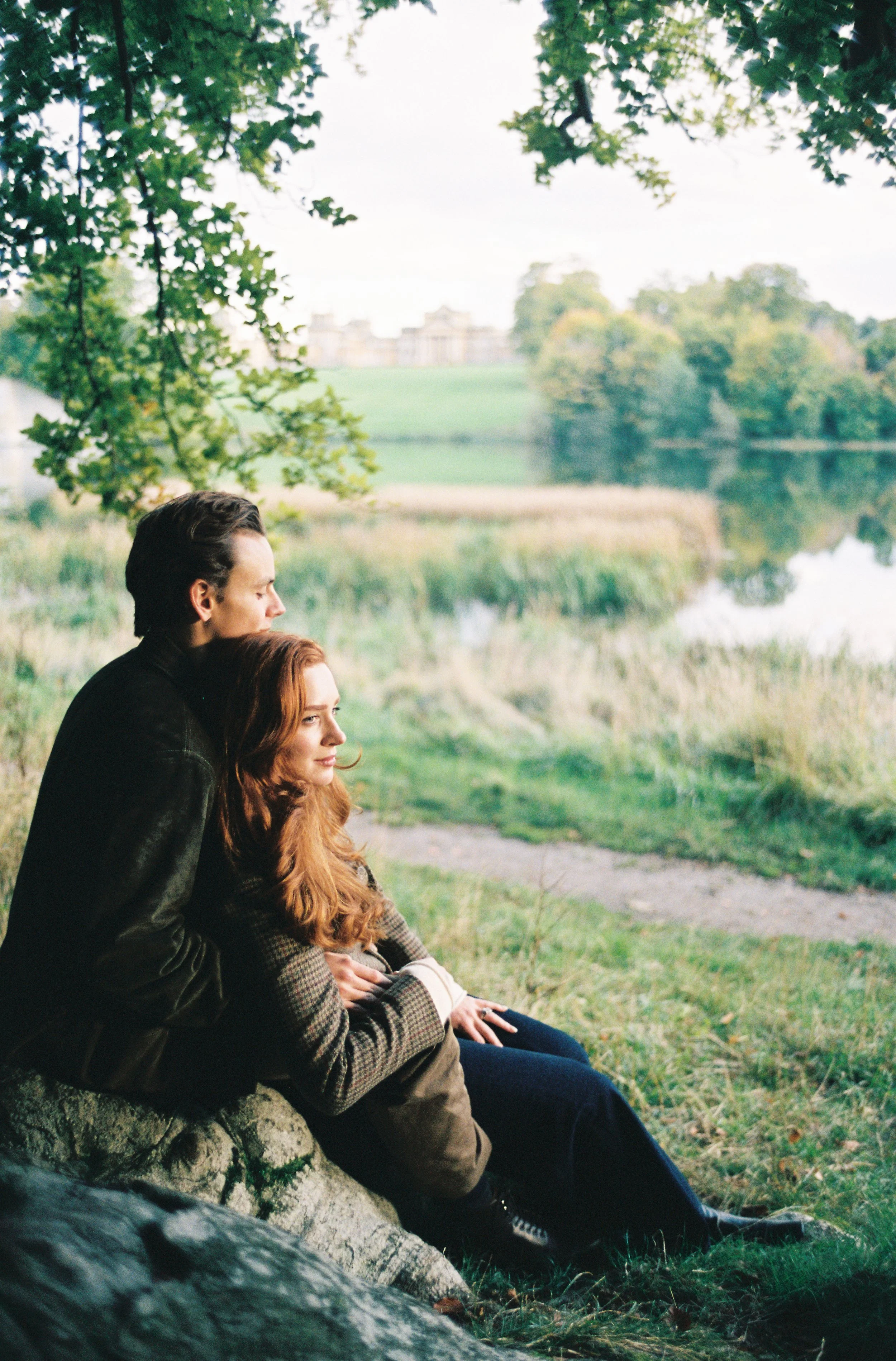 A young man and woman sit close together on a large rock near a pond, surrounded by grass and trees in a park, with a large building visible in the background.