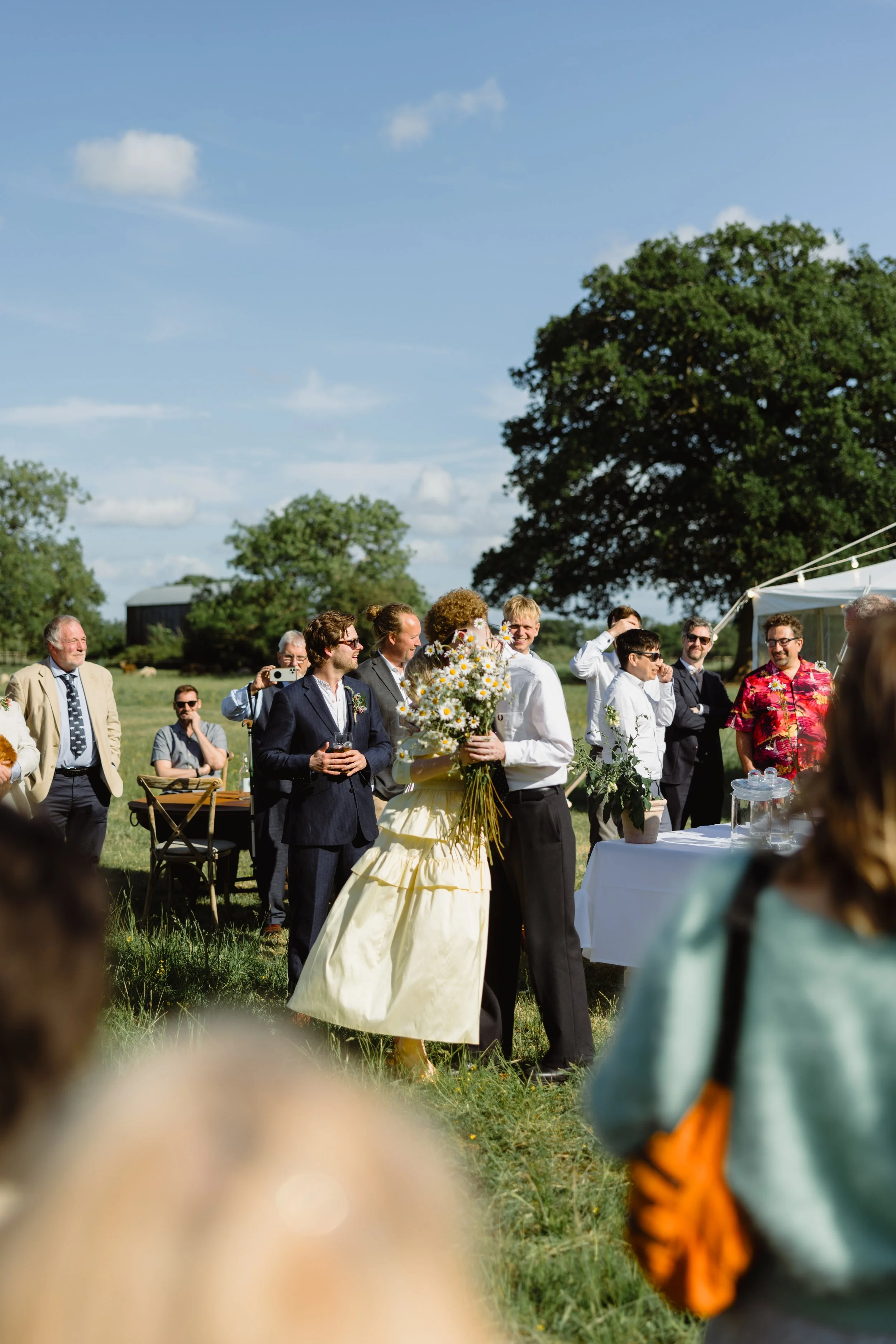 A wedding celebration outdoors with a bride in a yellow dress and a groom in a white shirt holding a bouquet of daisies, surrounded by guests on a sunny day with blue skies and large trees.
