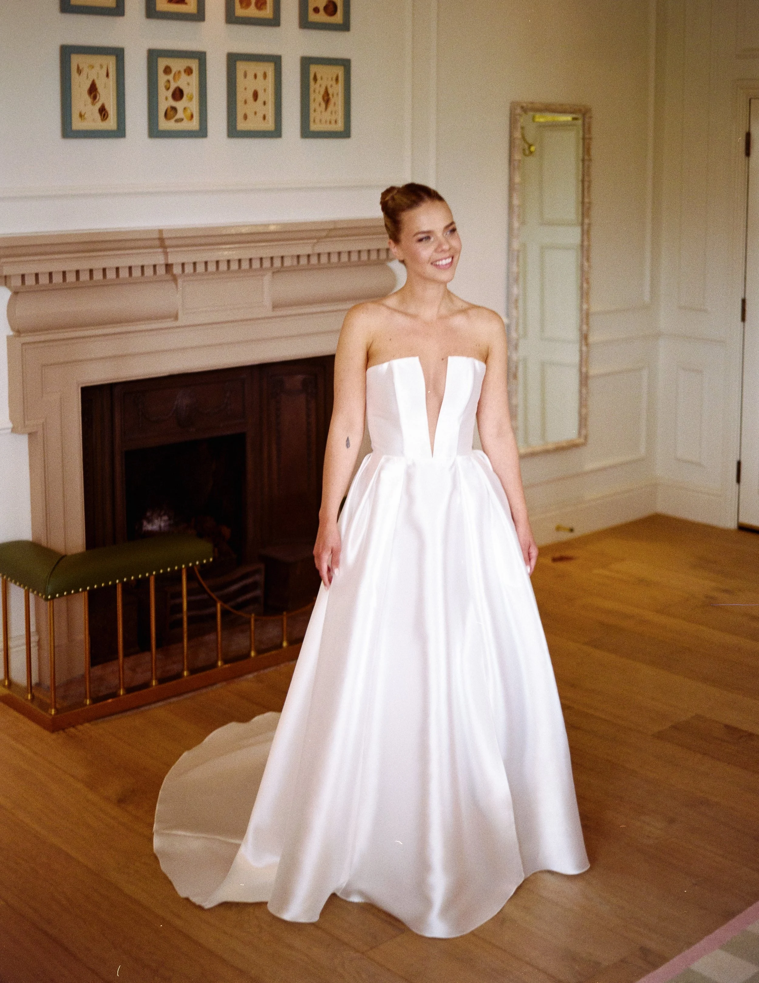A woman in a white strapless wedding dress with a deep V neckline standing in a room with wooden floors and white walls, smiling.