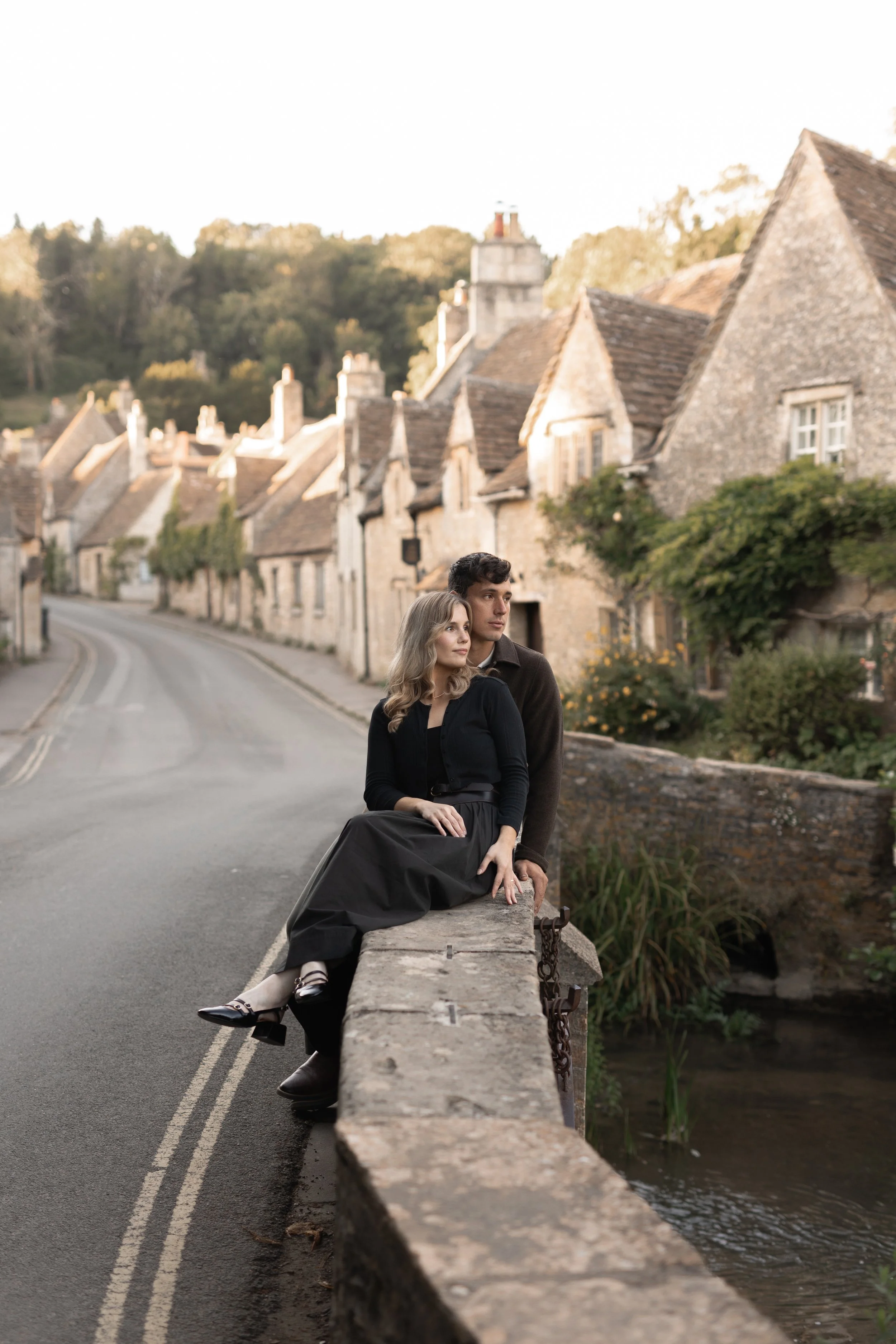 A man and woman sitting on a stone wall along a quiet, winding village street with traditional stone houses in the background. The woman is wearing a black dress and the man is in a dark sweater. The scene appears to be set during late afternoon or e