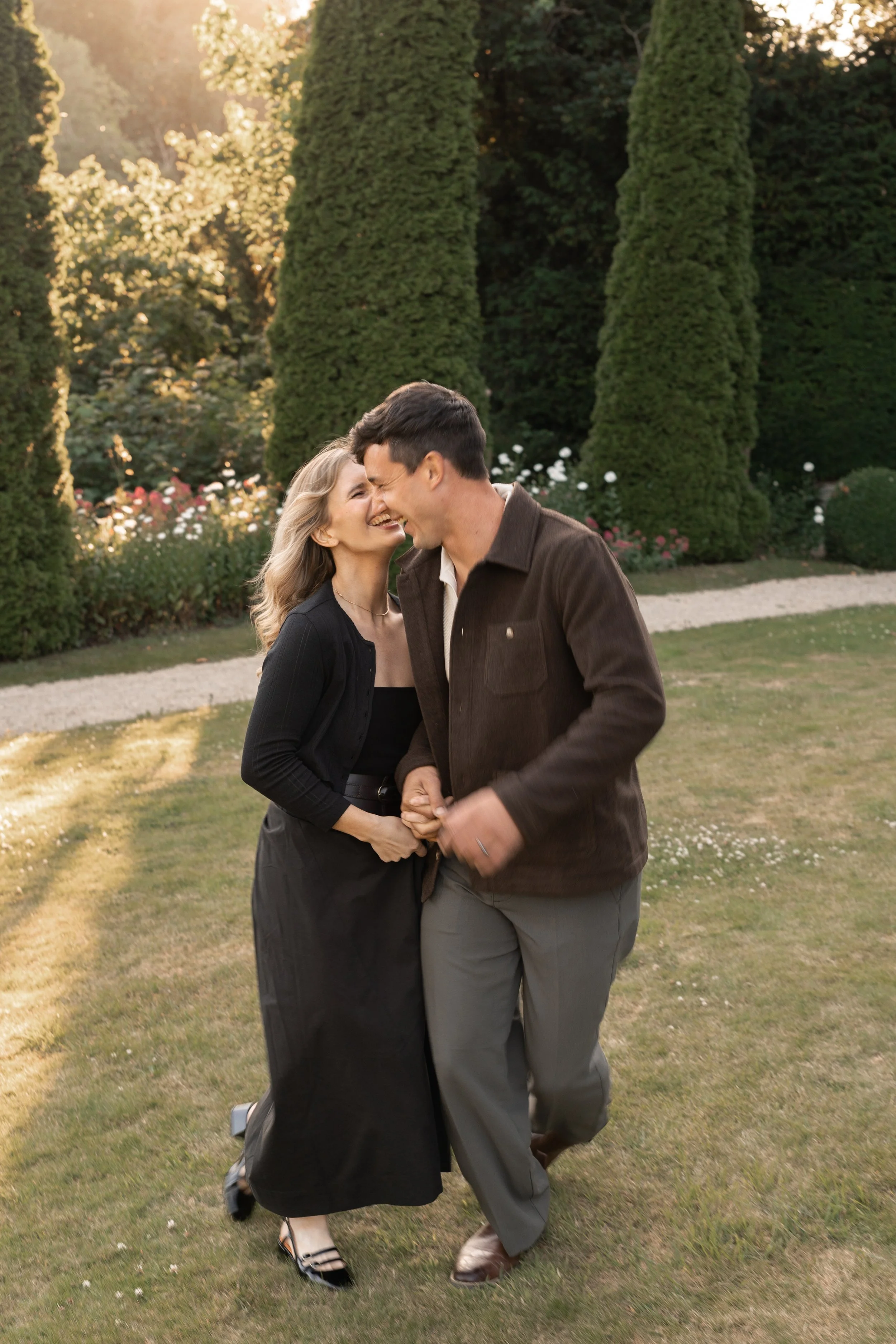 A woman and a man joyfully dancing outdoors in a garden with tall trees and flowers in the background, during sunset.
