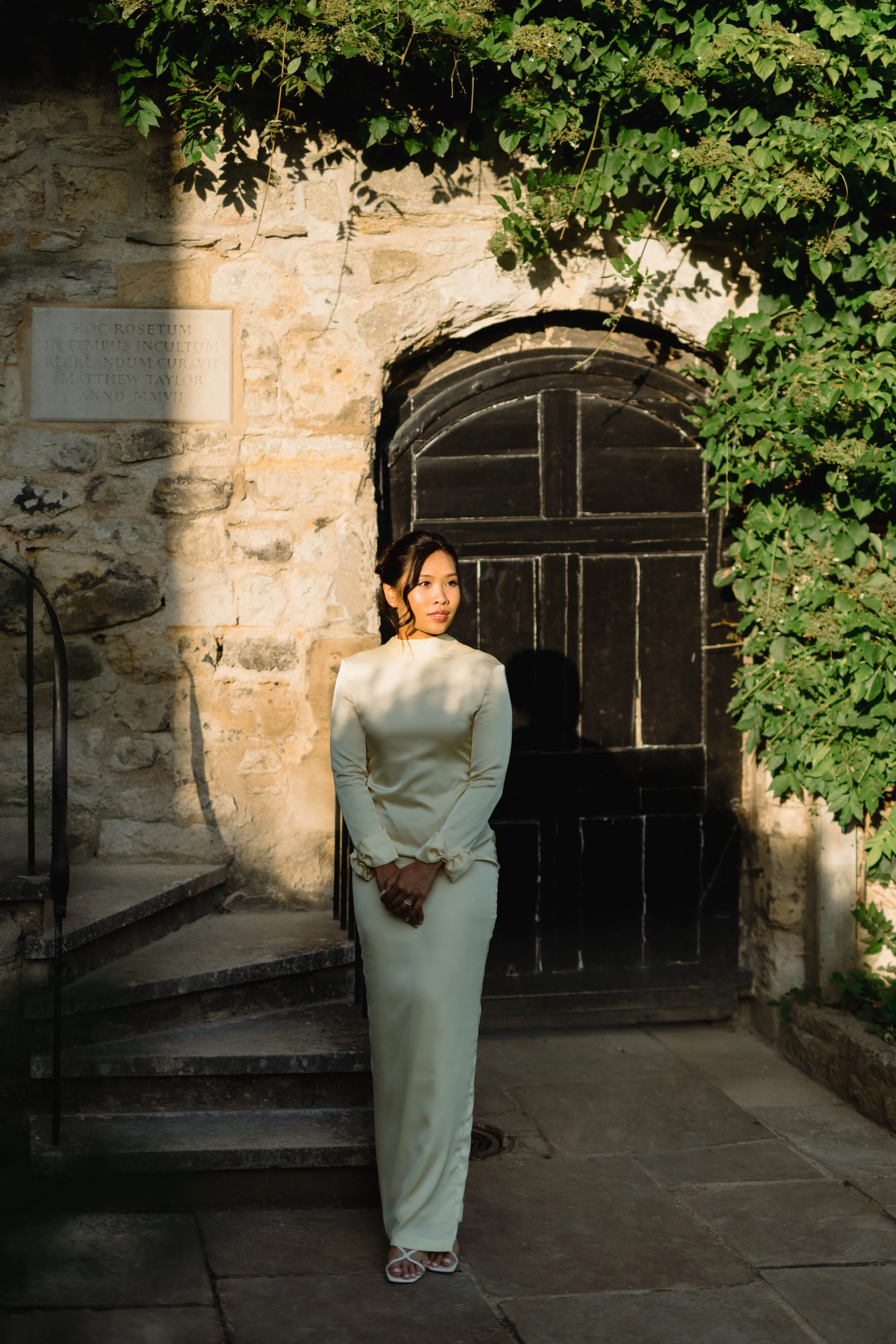 A woman in a long cream-colored dress standing on stone steps near a dark wooden door, surrounded by stone walls and green foliage, with sunlight casting shadows.