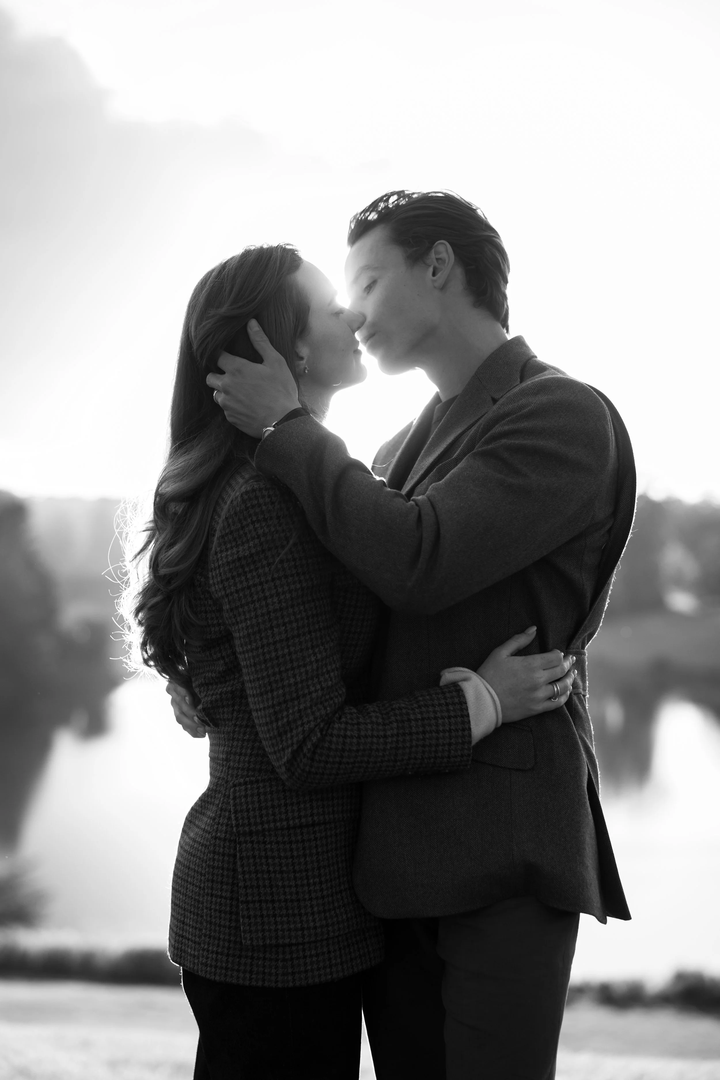 A black-and-white photo of a couple embracing outdoors at sunset, about to kiss, with water and trees in the background.