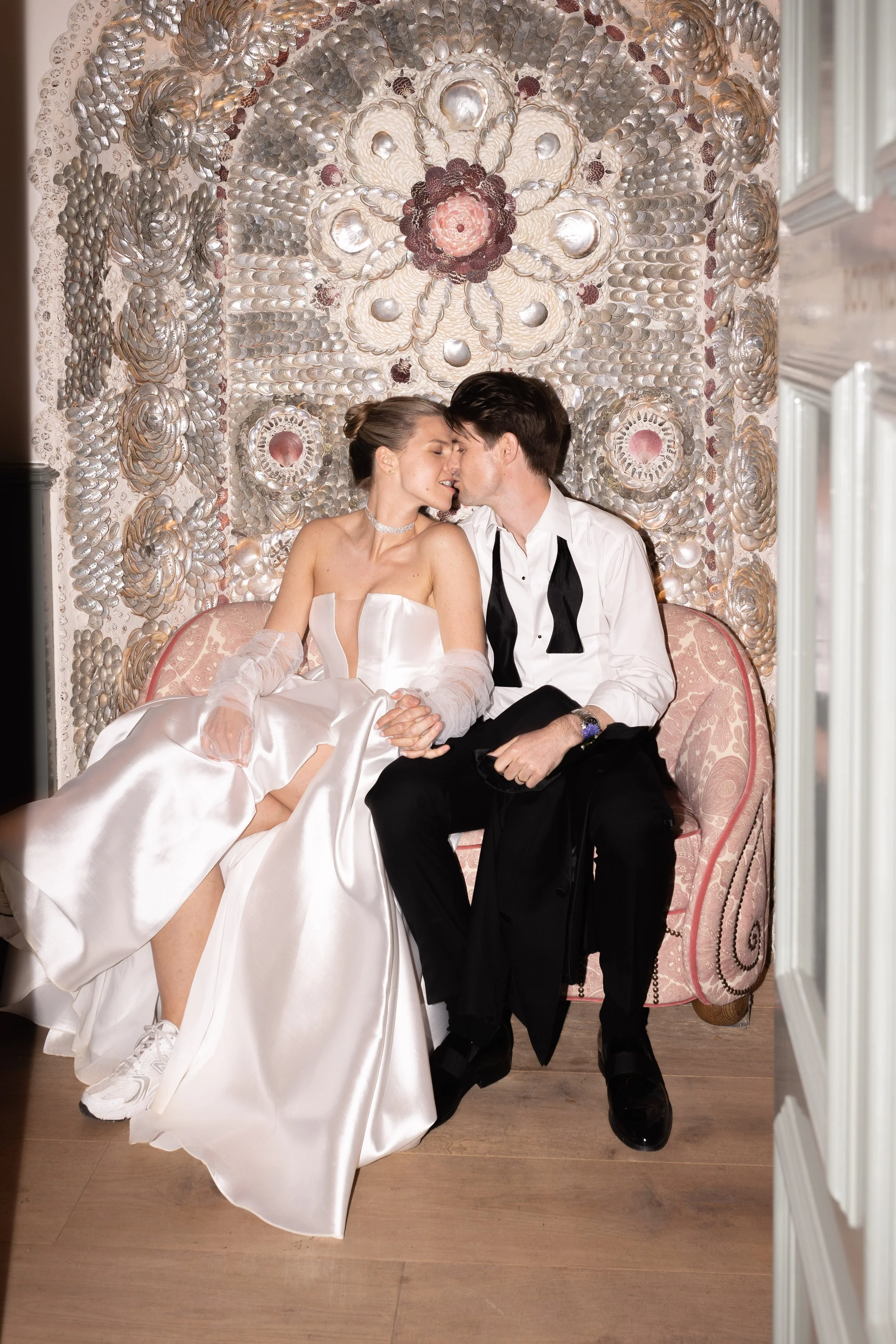 A bride and groom sitting closely on a pink vintage sofa, about to kiss, with a decorative shell mirror behind them.