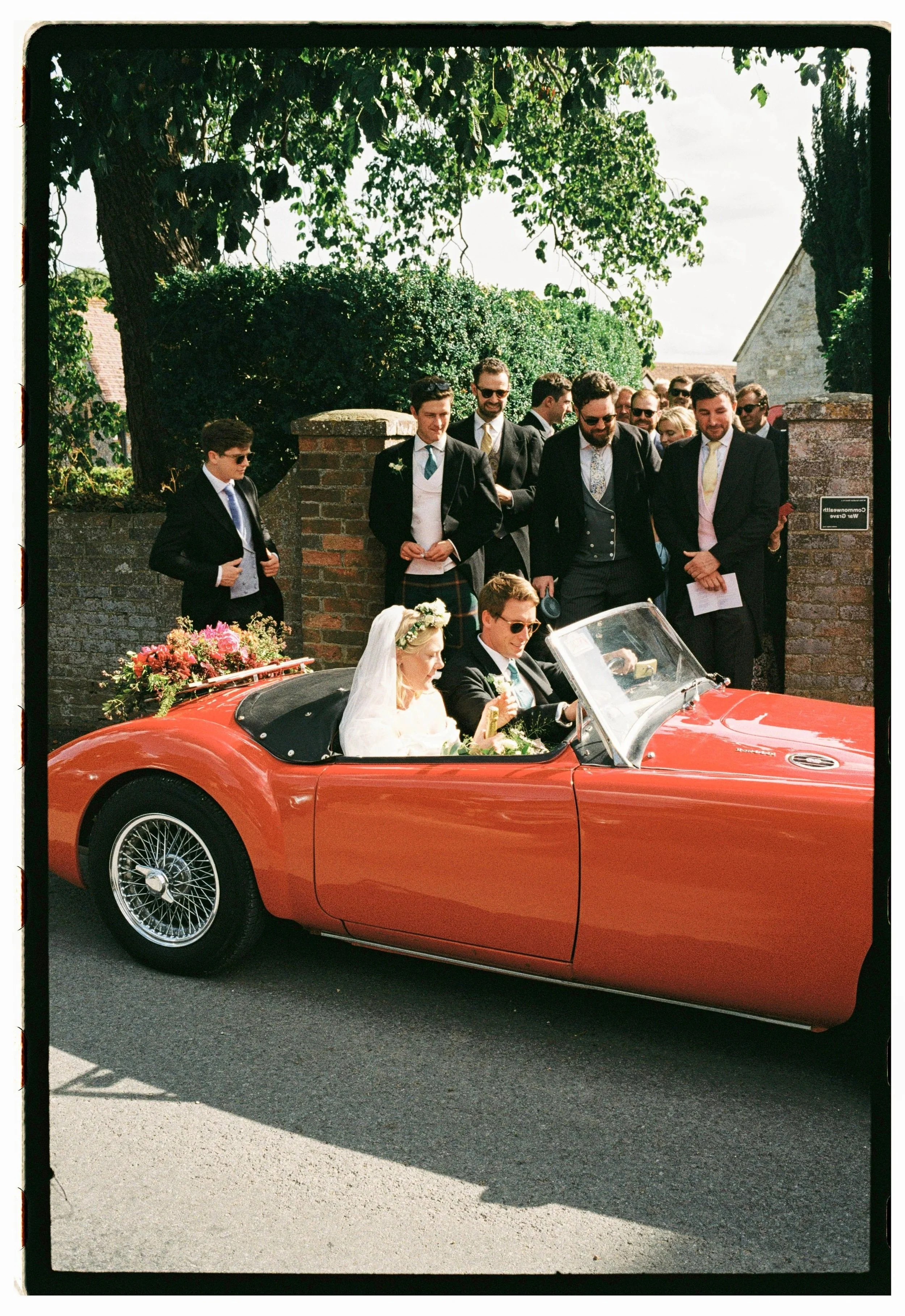 35mm bride and groom sitting in a vintage red convertible car, surrounded by friends in formal attire at a wedding celebration.