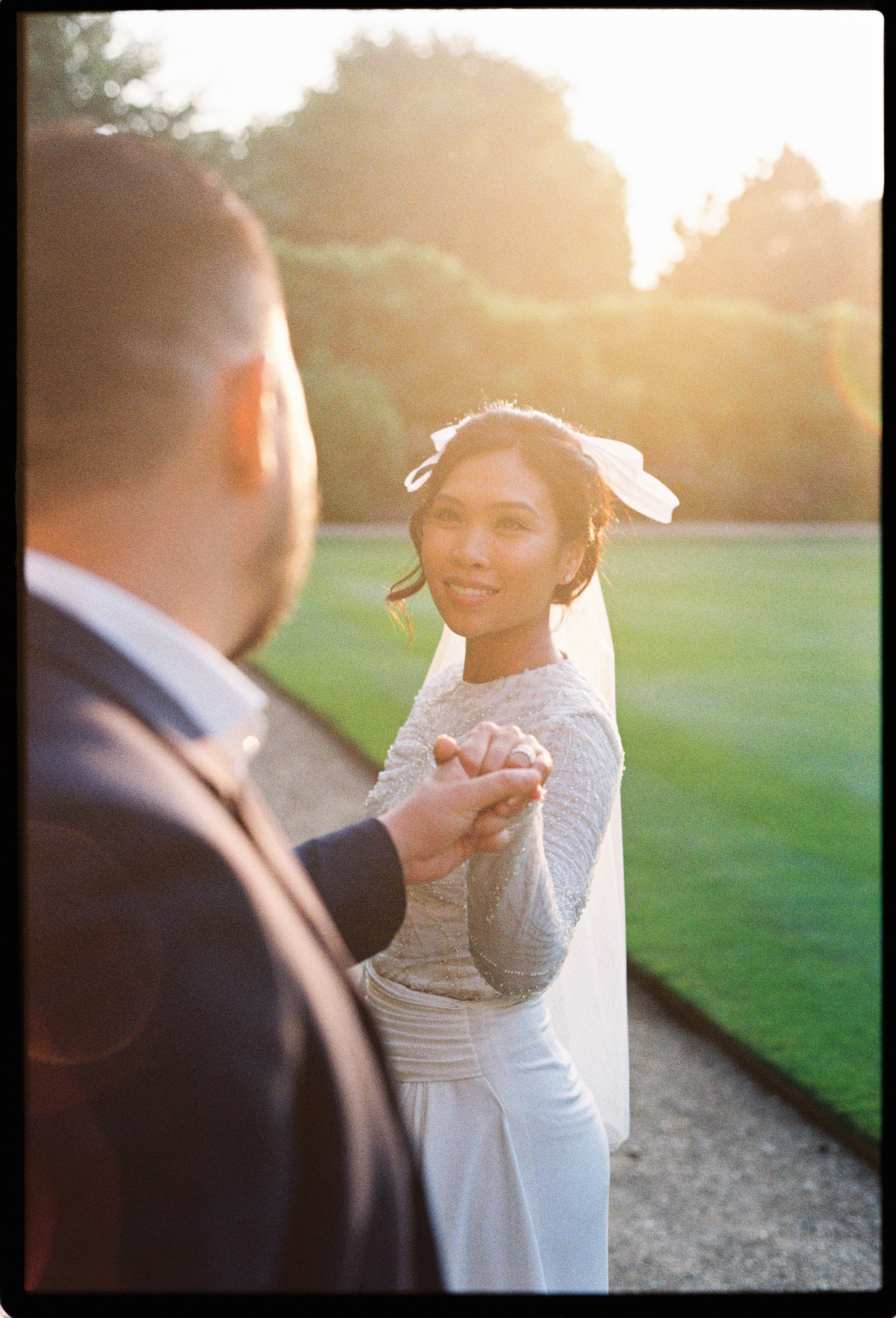A woman in a wedding dress holding hands with a man dressed in a suit outdoors during sunset.