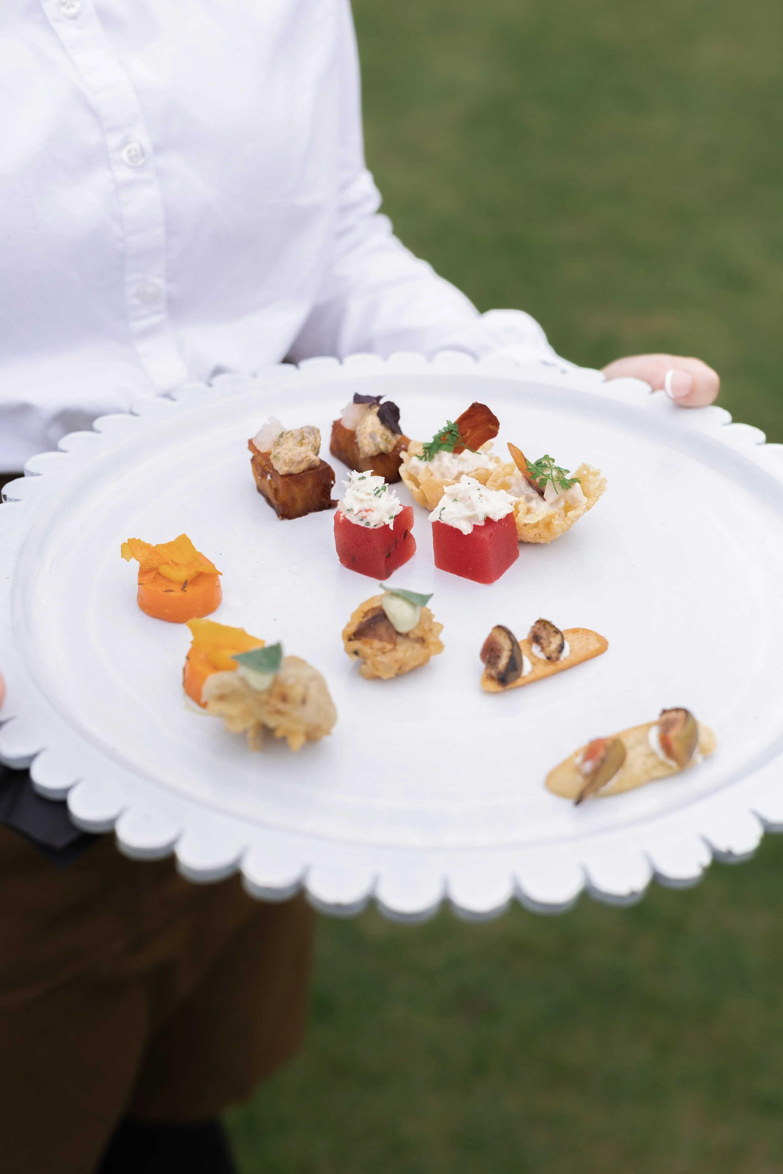 A person in a white shirt holding a white tray with various small, intricate appetizers or canapés on it.