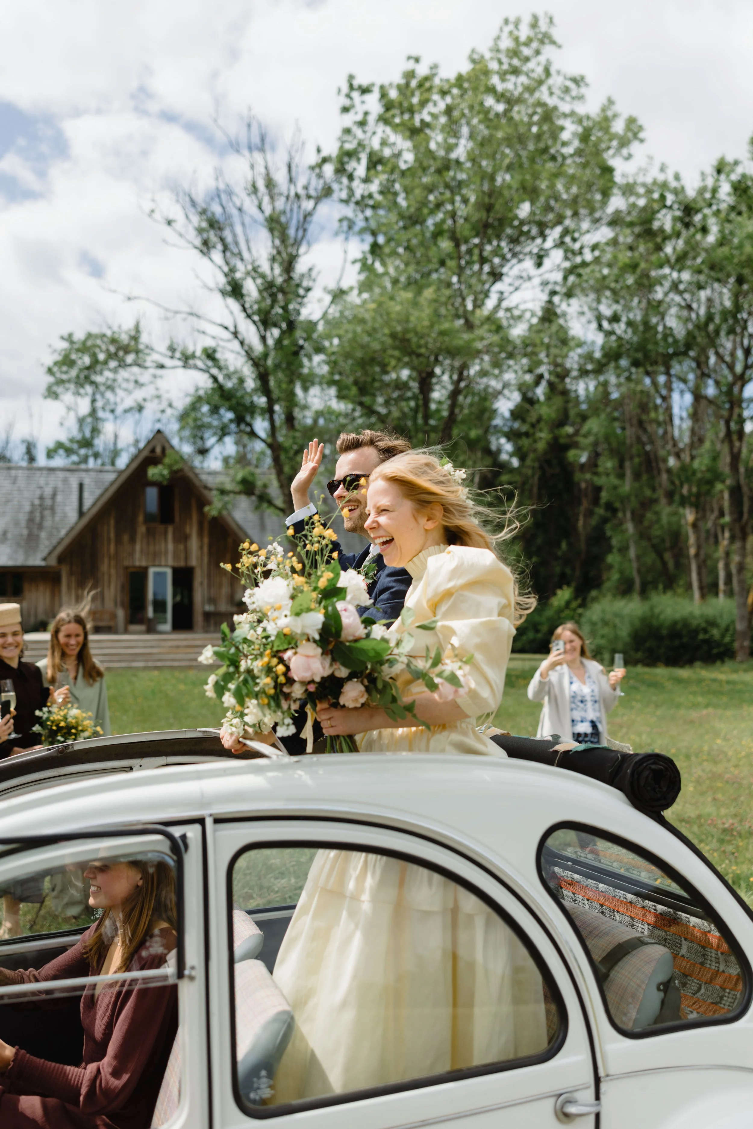 A bride and groom ride in a vintage car with a bride holding a bouquet, smiling and waving to guests who are taking photos and celebrating outside in a grassy area with trees and a wooden building in the background.